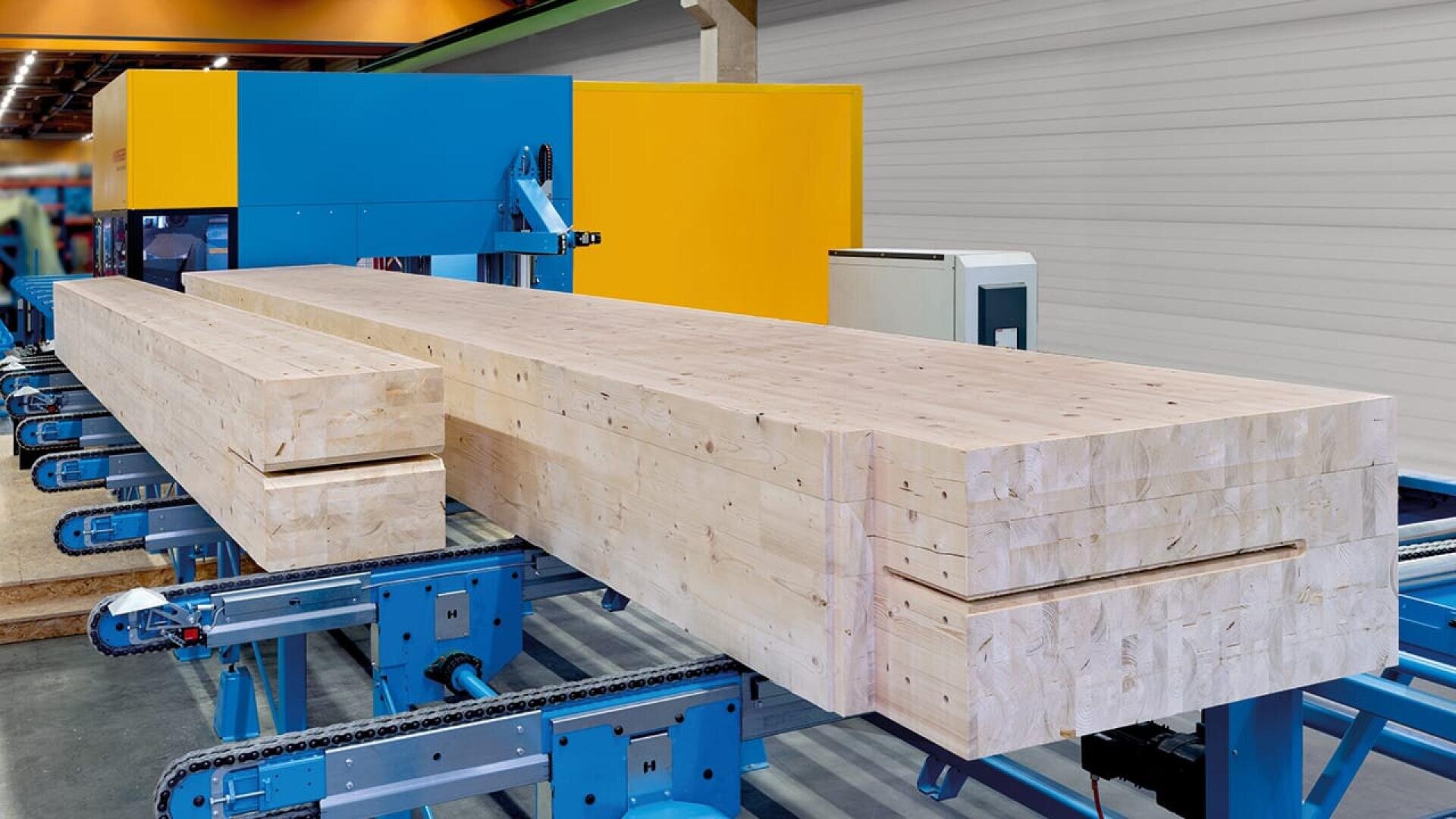 Eine große Industriemaschine bearbeitet und schneidet gestapelte Holzbalken in einer Fabrikumgebung, wobei blaue Fördersysteme das Holz tragen.