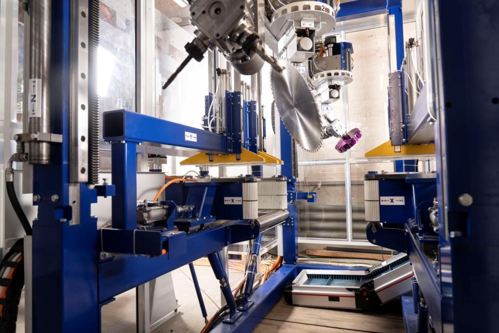 Nahaufnahme einer industriellen Automatisierungsausrüstung, die aus blauen Metallrahmen, Roboterarmen, Kabeln und Maschinenteilen in einer Fabrik besteht. Das Licht dringt von links ein und hebt die mechanischen Komponenten hervor.