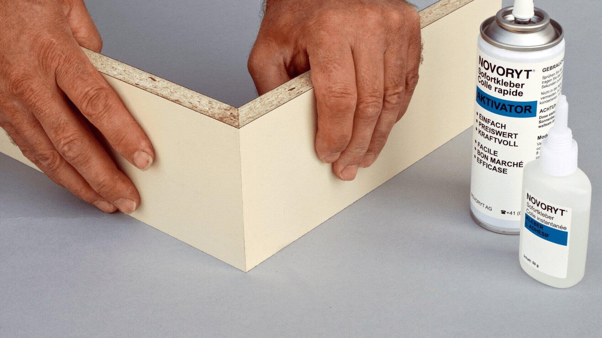 Zwei Hände fügen zwei Holzstücke in einem rechten Winkel zusammen. In der Nähe befinden sich eine Dose mit der Aufschrift "Activator" und eine kleine Flasche, die wahrscheinlich beide als Klebstoff für die Holzbearbeitung verwendet werden.