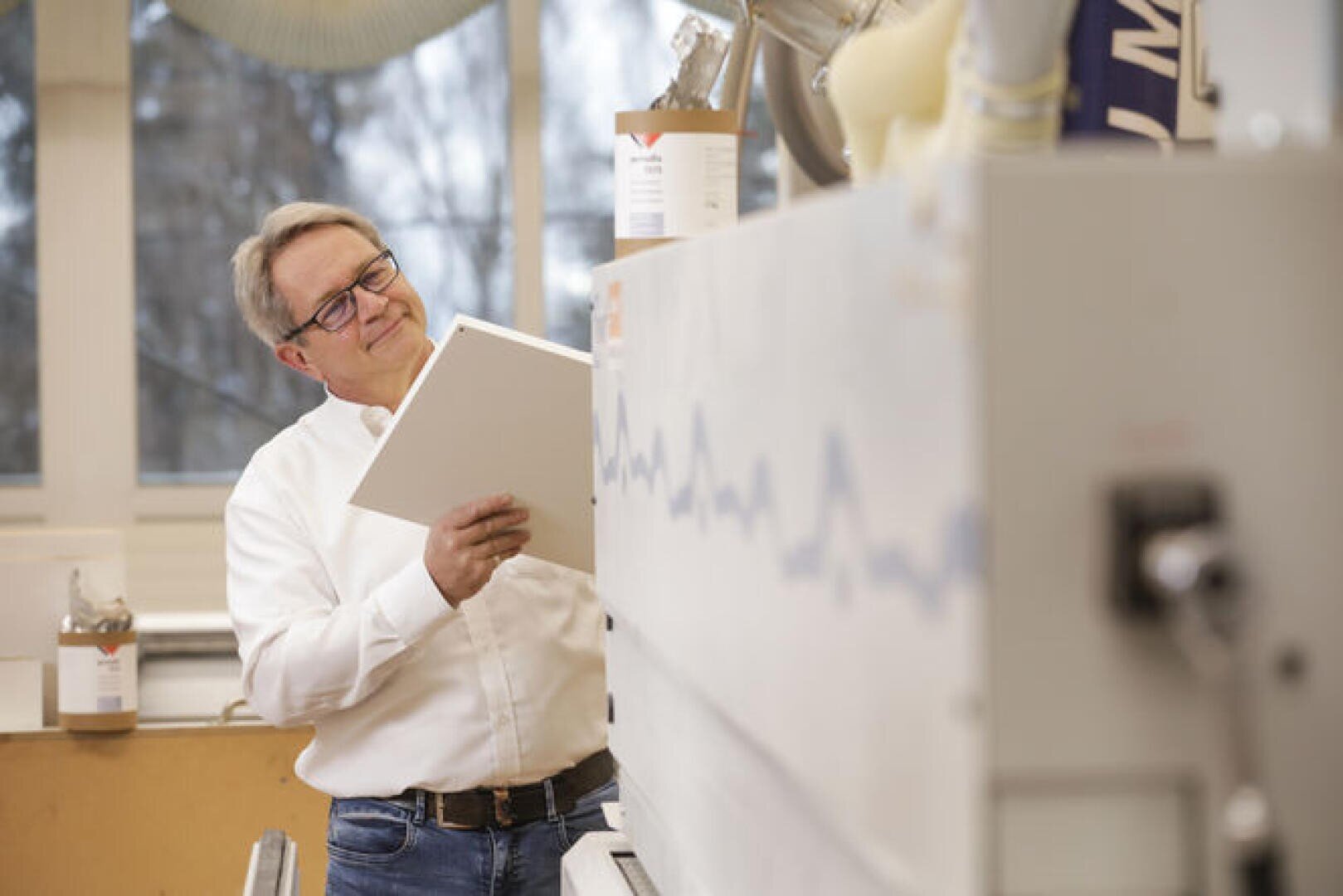 Un homme portant des lunettes et une chemise blanche se tient dans un laboratoire lumineux, tenant un dossier et regardant une grande machine scientifique avec un motif d'ondes bleues. Des équipements de laboratoire sont visibles à l'arrière-plan.