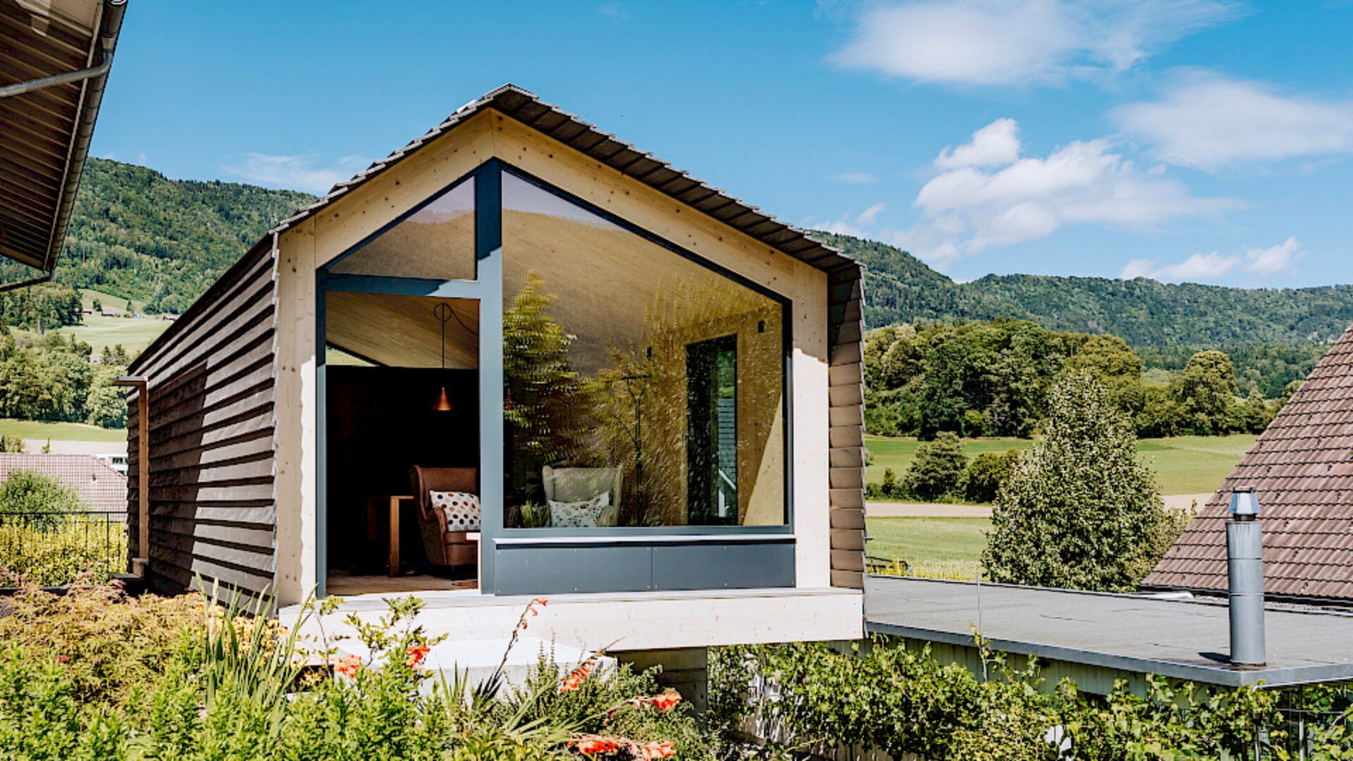 Moderne Holzhütte mit großen Glasfenstern mit Blick auf eine üppig grüne Landschaft und Hügel unter einem blauen Himmel mit vereinzelten Wolken; im Inneren der Hütte ist ein Teil eines Sofas zu sehen.