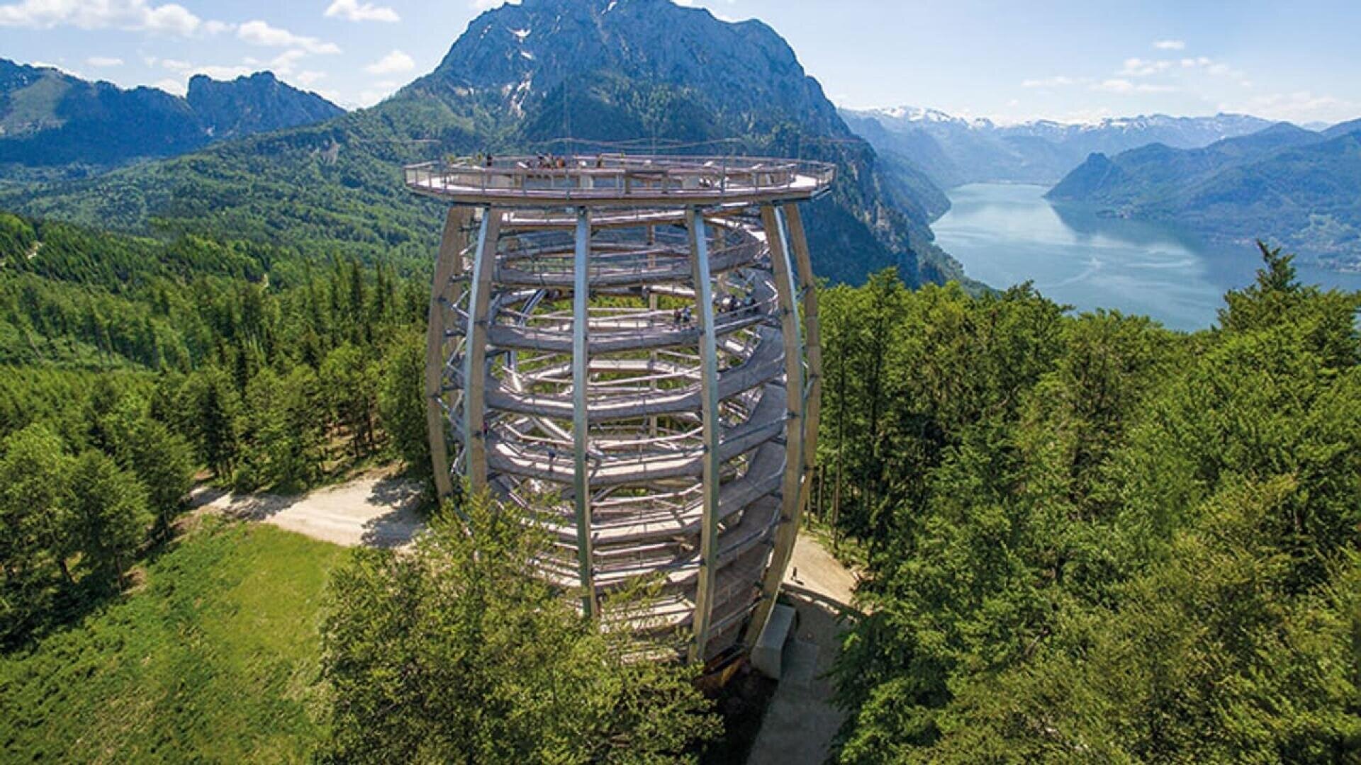 Une grande tour d'observation en bois avec une rampe en spirale s'élève au-dessus d'une forêt, offrant une vue panoramique sur les montagnes environnantes, un lac sinueux et une verdure luxuriante sous un ciel bleu clair.