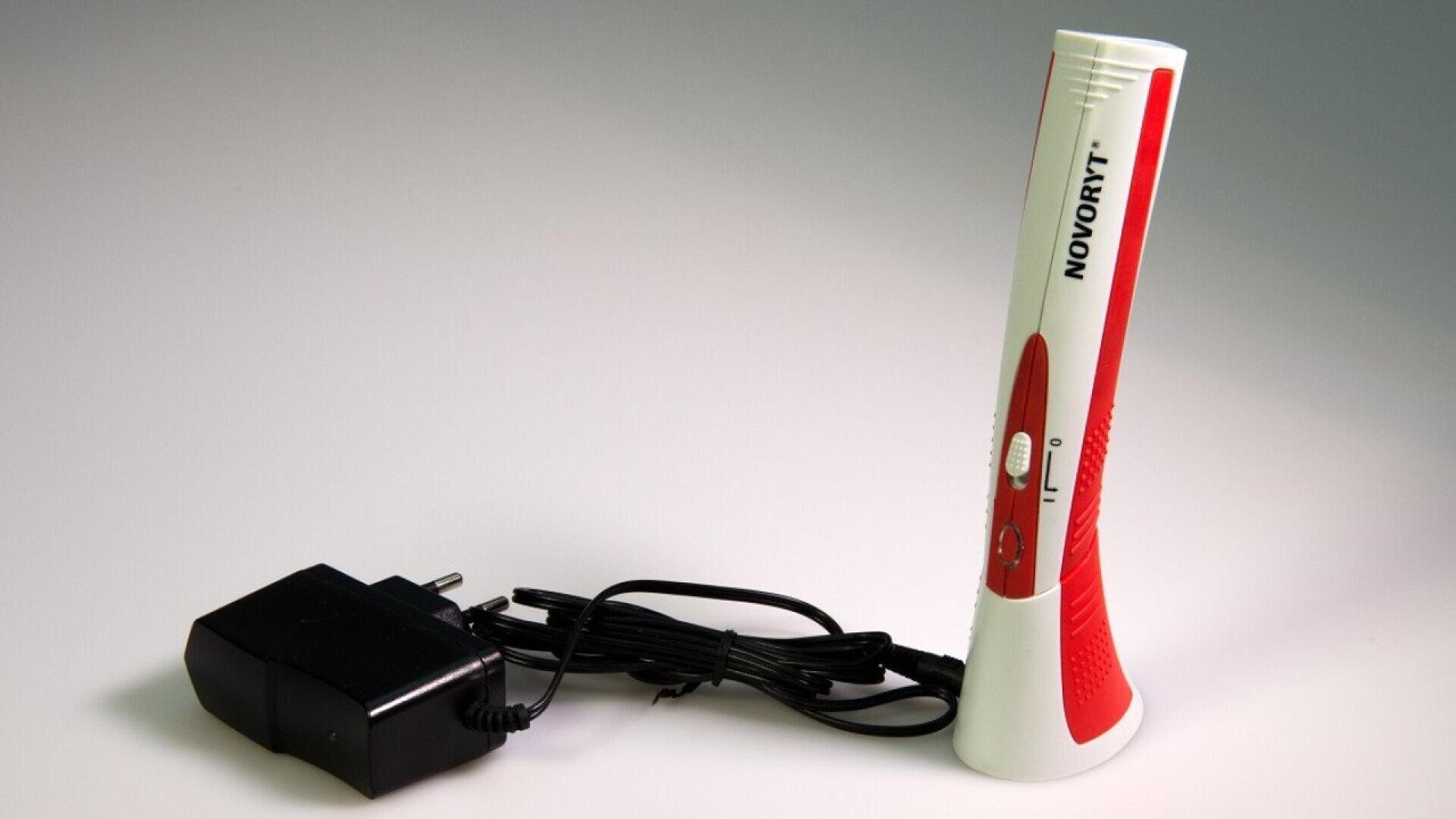 Un appareil électronique portable rouge et blanc étiqueté Novoryt se tient debout à côté d'un adaptateur d'alimentation noir avec un cordon connecté sur un fond clair.