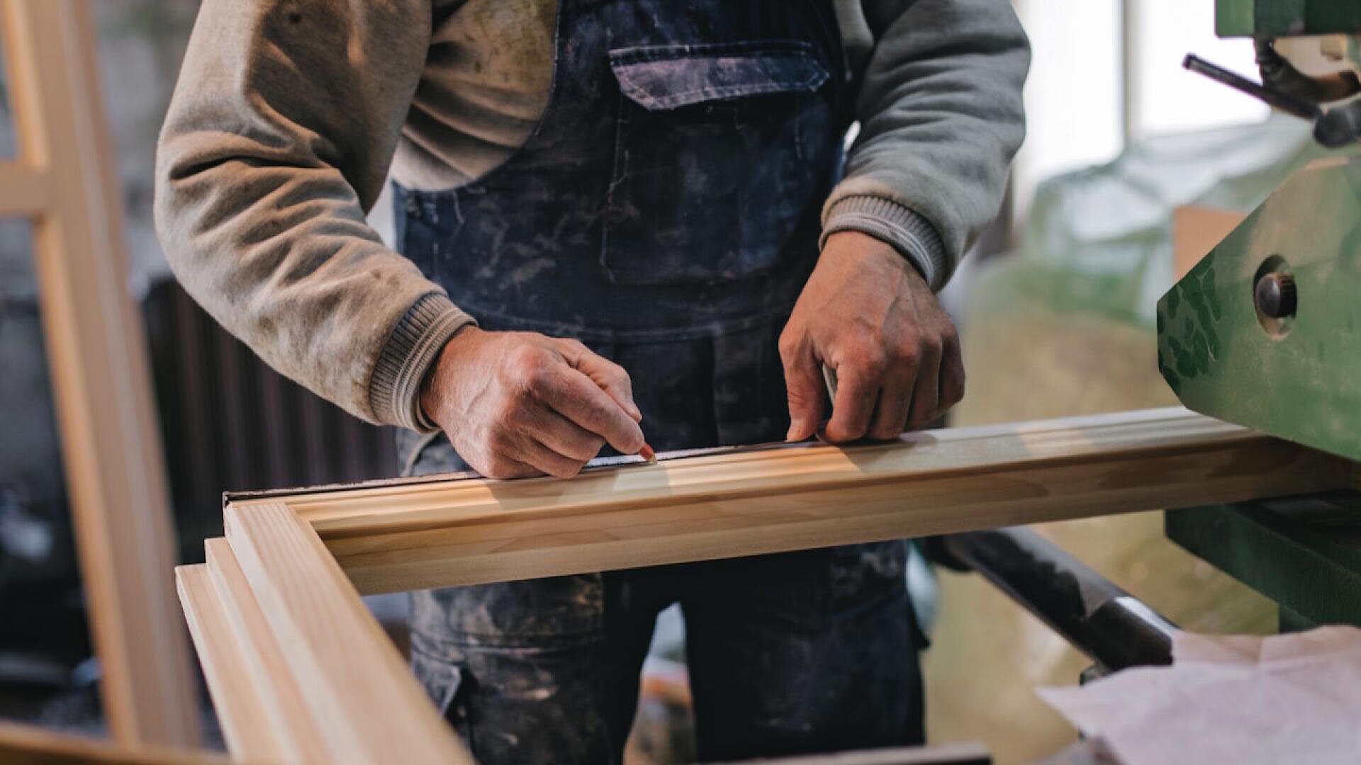 Une personne en combinaison de travail mesure et marque un cadre en bois avec un crayon dans un atelier afin de préparer le bois pour la découpe ou le montage.