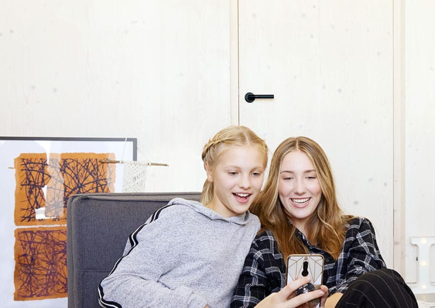 Zwei lächelnde Mädchen sitzen auf einer Couch und schauen gemeinsam auf ein Smartphone. Die eine trägt einen grauen Kapuzenpulli, die andere ein kariertes Hemd. Sie scheinen glücklich und beschäftigt zu sein. Im Hintergrund ist ein gerahmtes abstraktes Kunstwerk zu sehen.