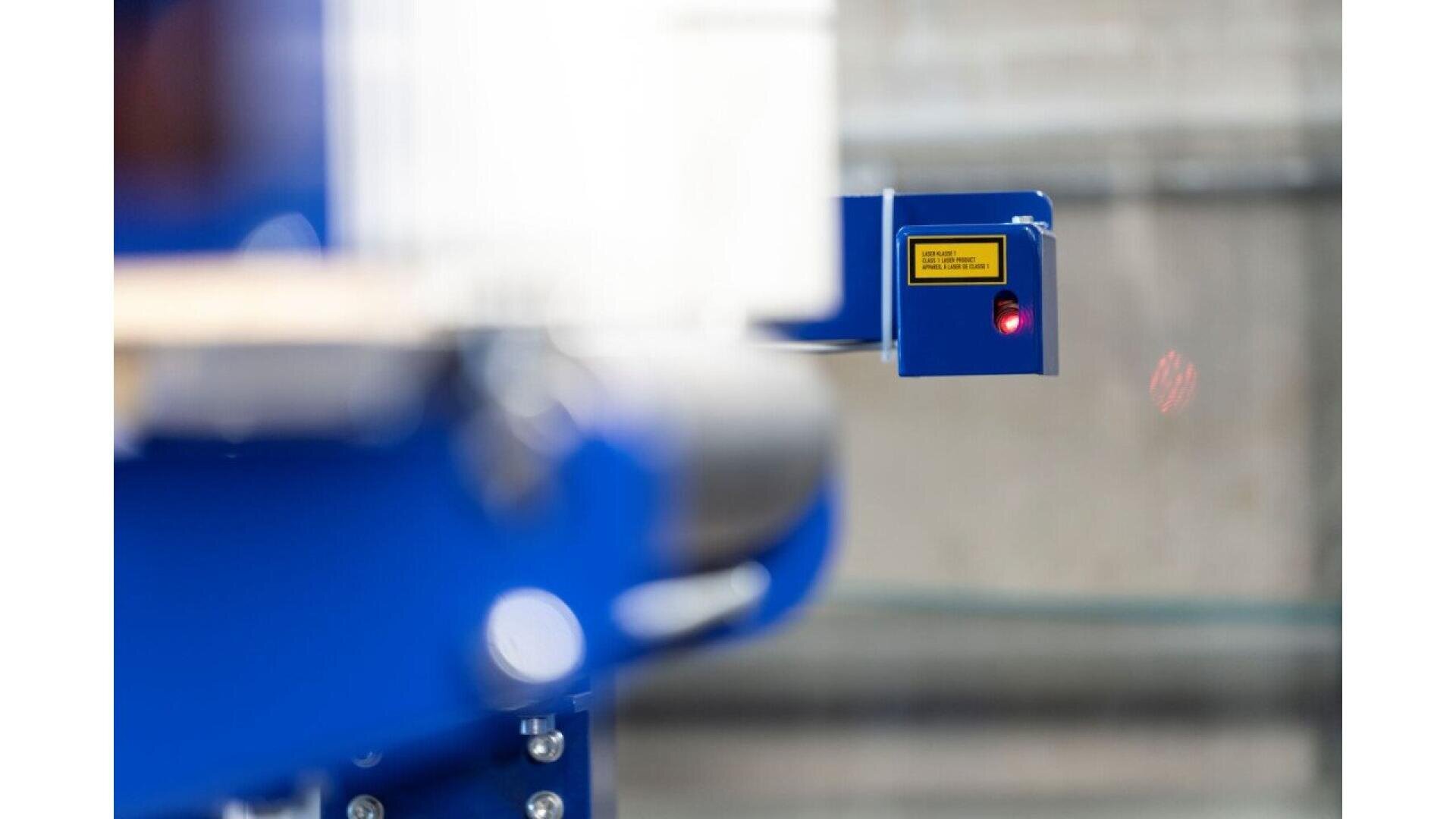 Gros plan d'un capteur industriel bleu avec une étiquette d'avertissement jaune et un voyant rouge, monté sur une machine, avec un arrière-plan flou.