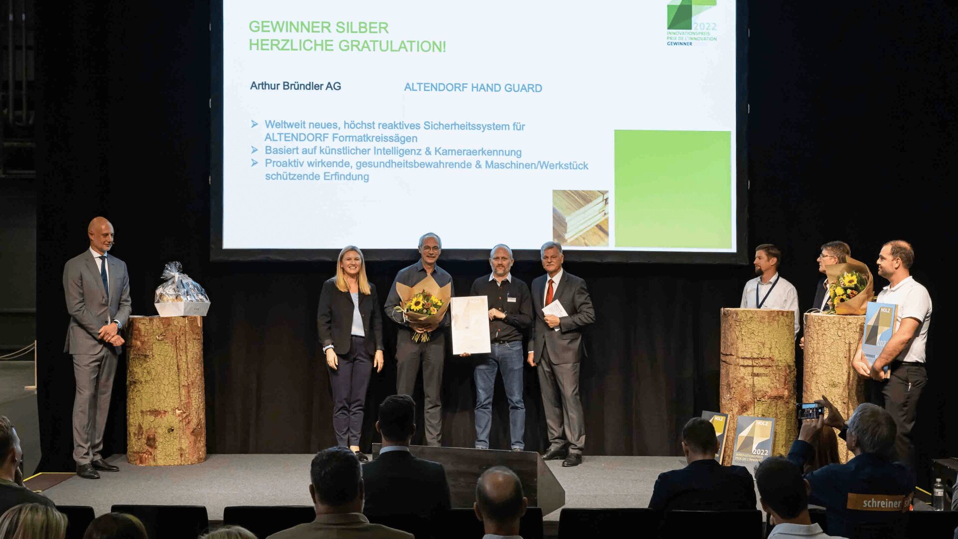 Eine Gruppe von Personen steht bei einer Preisverleihung auf der Bühne. Eine Person hält Blumen, eine andere eine Urkunde in der Hand. Eine Präsentationsfolie hinter ihnen kündigt GEWINNER SILBER an und nennt Einzelheiten über das ausgezeichnete Projekt.