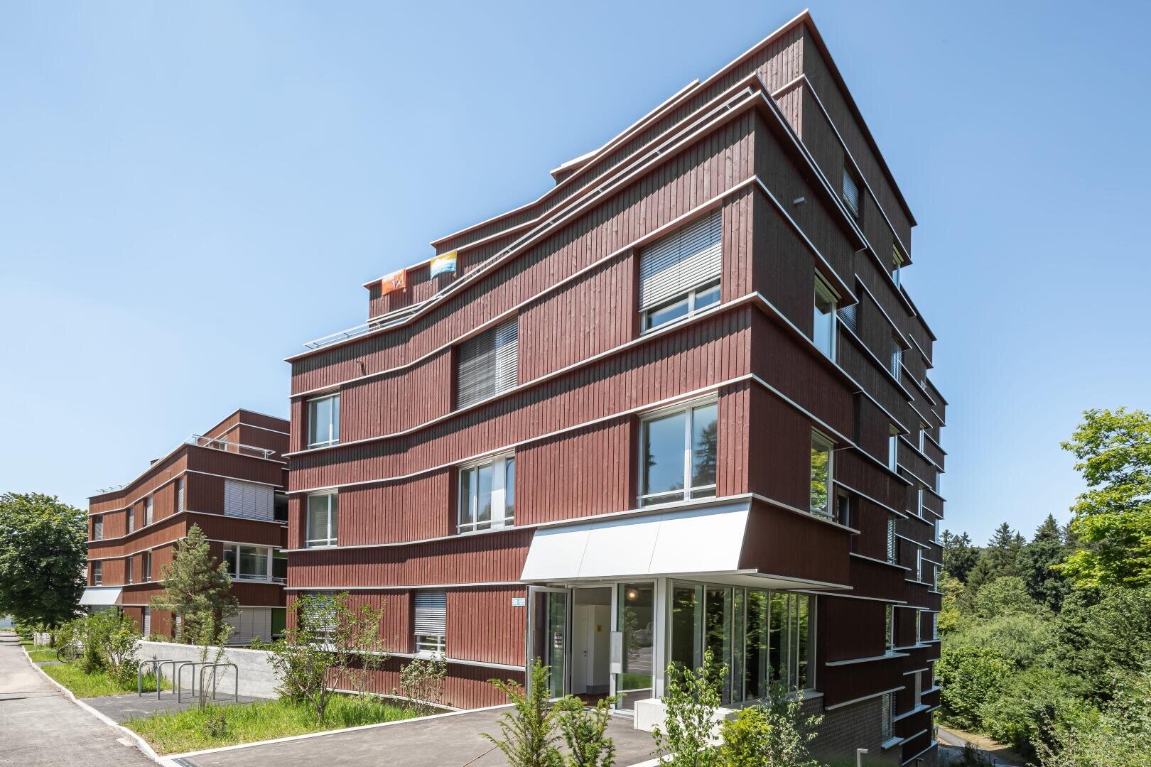 Ein modernes mehrstöckiges Wohnhaus mit roter Holzfassade, großen Fenstern und weißen Akzenten in einer grünen, baumbestandenen Gegend unter einem klaren blauen Himmel.