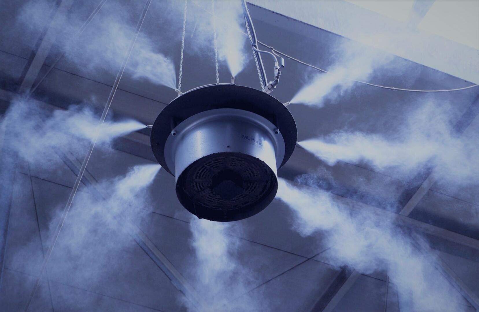 Ein großes, kreisförmiges, an der Decke montiertes Gerät gibt Nebel- oder Dampfströme in mehrere Richtungen in einer industriellen oder lagerähnlichen Umgebung mit freiliegenden Trägern ab.