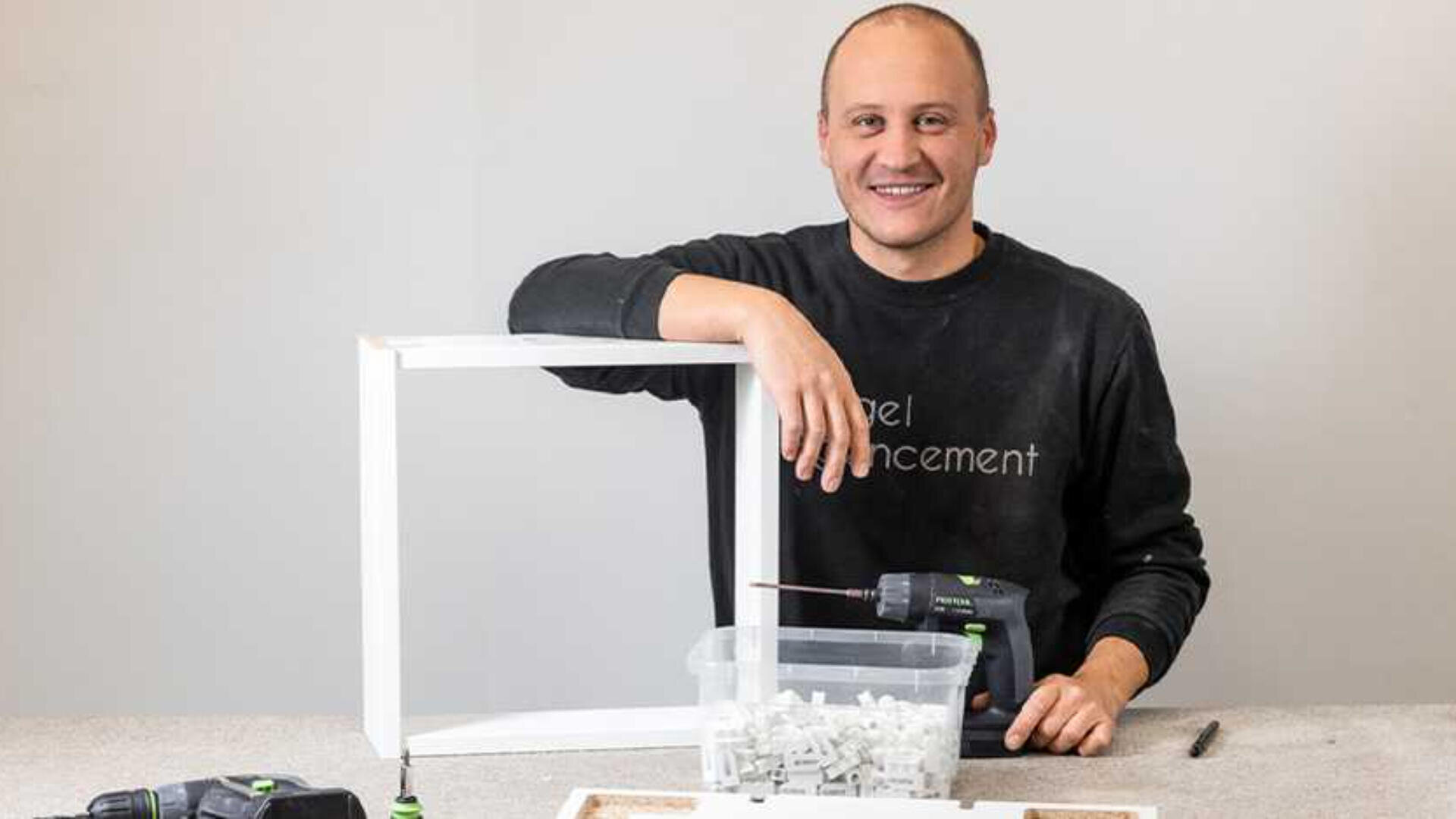 Eine lächelnde Person in einem schwarzen Hemd steht hinter einem weißen rechteckigen Rahmen und hält eine Bohrmaschine in der Hand. Ein Plastikbehälter mit weißen Plastikteilen und eine weitere Bohrmaschine stehen auf dem Tisch vor ihnen.