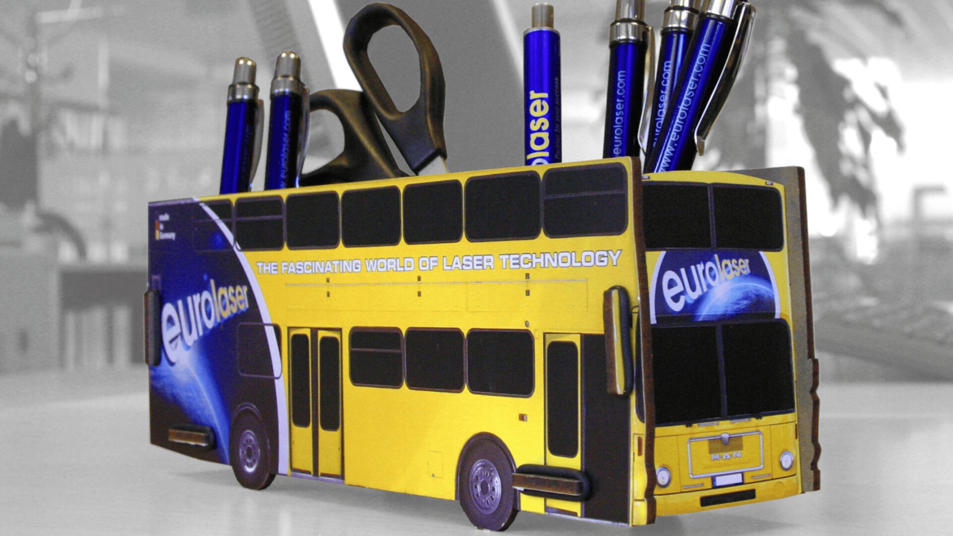 Ein gelber Doppeldeckerbus in Form eines Stifthalters mit der Aufschrift eurolaser hält blaue Stifte und eine Schere auf einem Schreibtisch in einer Büroumgebung.
