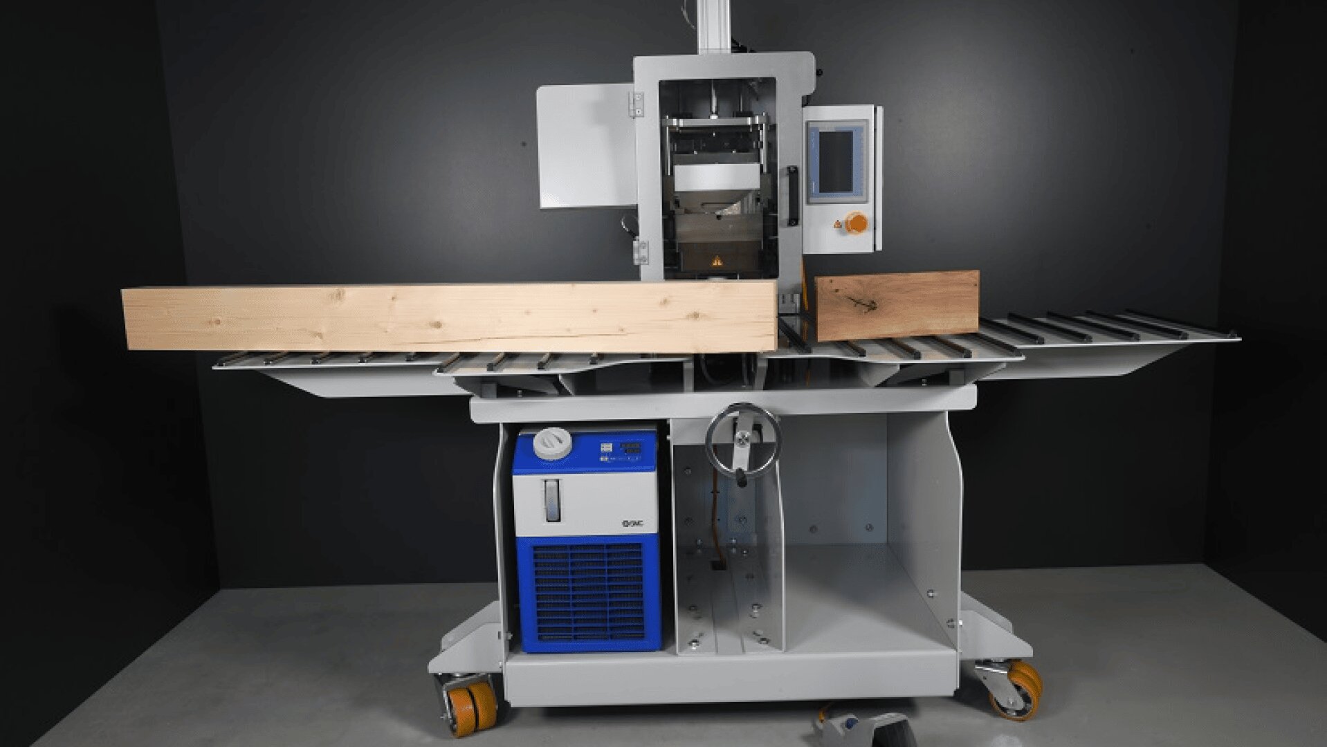 Eine Holzbearbeitungsmaschine mit Metallrollen hält zwei Holzbretter; sie verfügt über ein digitales Bedienfeld und ein blaues Kühlaggregat am Sockel, vor einem dunklen Hintergrund.