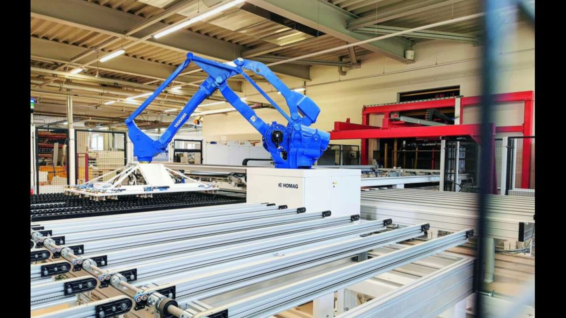 Ein blauer Industrieroboterarm arbeitet über Förderbändern in einer modernen Fabrik, umgeben von Metallgerüsten und Maschinen unter einer hohen Decke mit freiliegenden Balken.