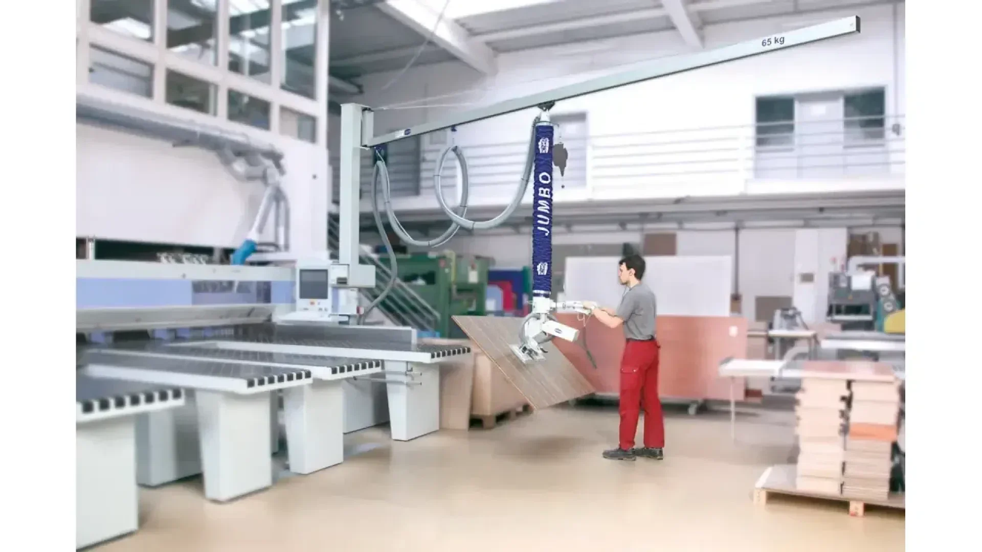 Un ouvrier en pantalon rouge utilise un appareil de levage industriel bleu pour déplacer une grande planche en bois dans une usine moderne et lumineuse, avec des machines et des tables de travail en arrière-plan.