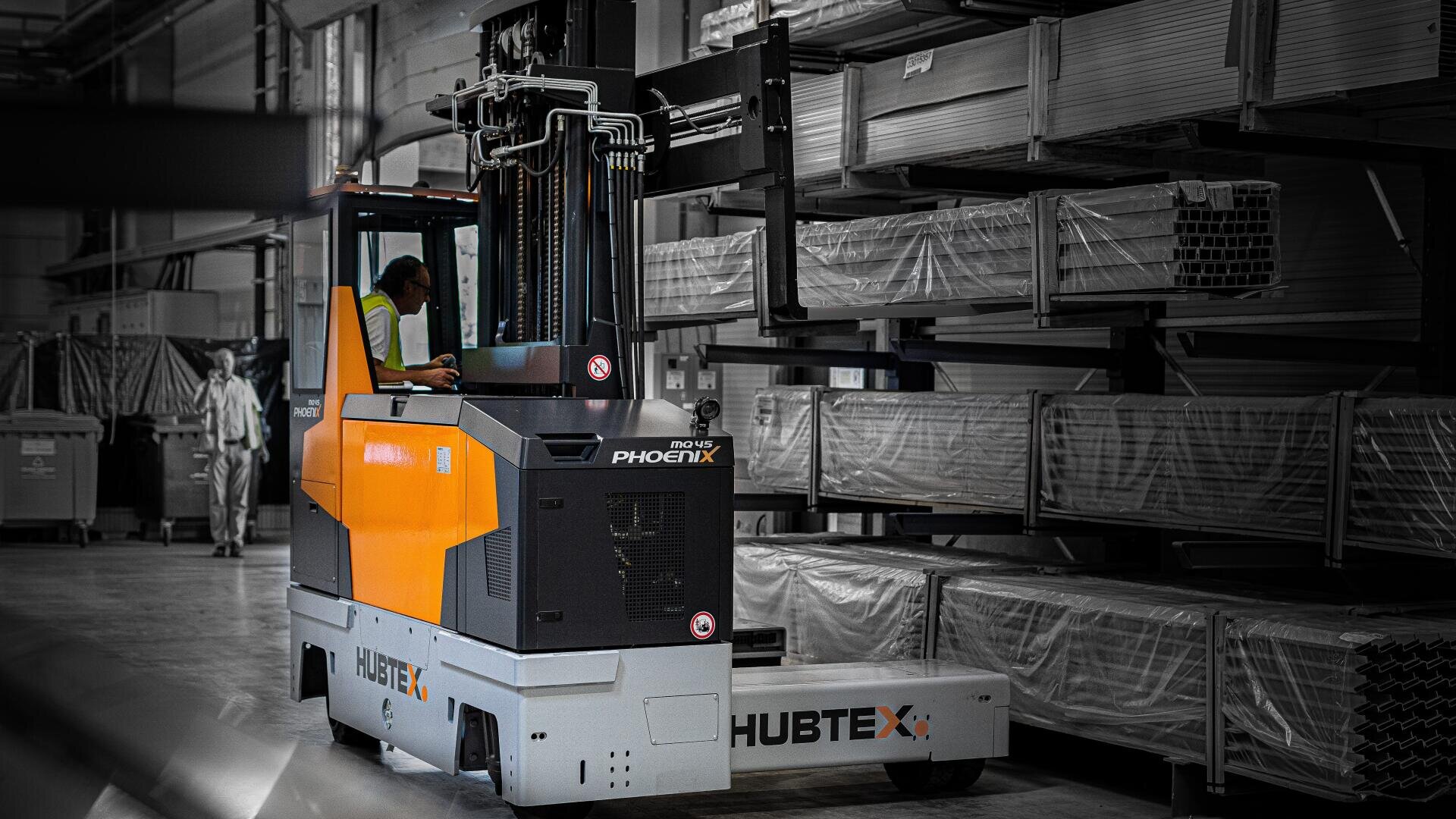 Ein Arbeiter bedient einen HUBTEX PHOENIX-Gabelstapler und hebt verpackte Bleche in einem Industrielager mit hohen Lagerregalen. Im Hintergrund sieht man eine weitere Person gehen.