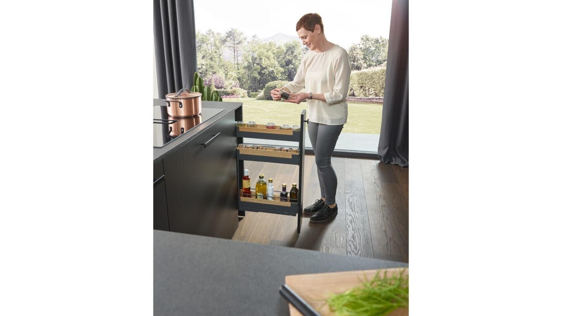 Une personne se tient dans une cuisine moderne à côté d'un îlot, rangeant de petits bocaux sur une étagère à épices coulissante. De grandes fenêtres et une vue sur une cour verdoyante sont visibles à l'arrière-plan.