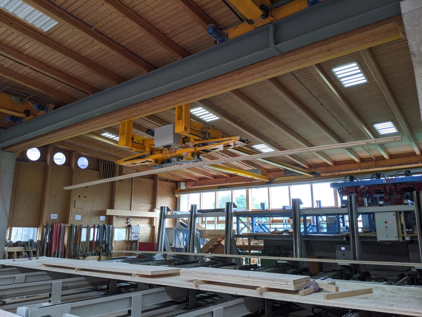 Eine große Industriehalle mit Holzbalken, Stahlbaumaterial und einem gelben Kransystem, das eine Holzplatte anhebt. Natürliches Licht dringt durch die Fenster und Lichtschächte ein. Stapel von Materialien sind im Arbeitsbereich sichtbar.