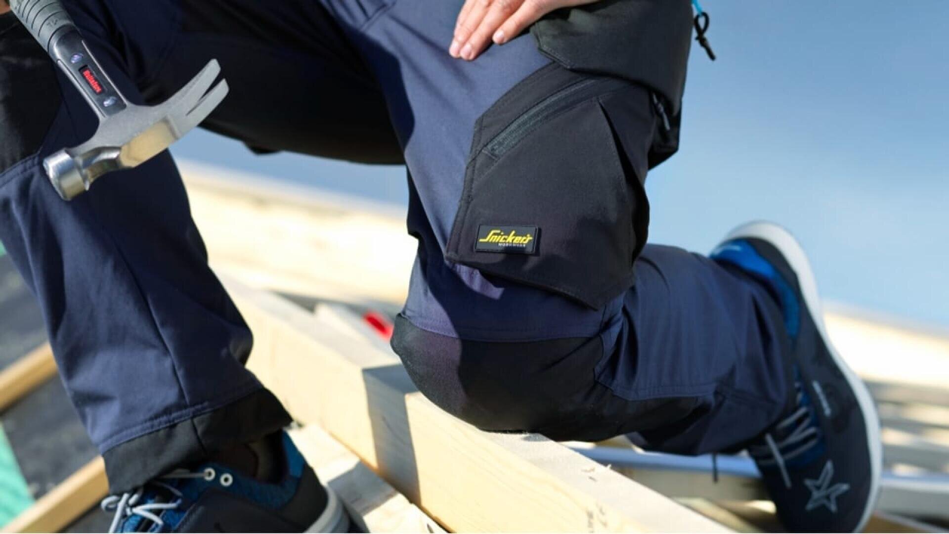 Une personne portant un pantalon de travail bleu marine et des baskets est agenouillée sur une charpente en bois et tient un marteau. Le pantalon porte un logo Snickers Workwear jaune sur la poche. Le ciel est dégagé à l'arrière-plan.