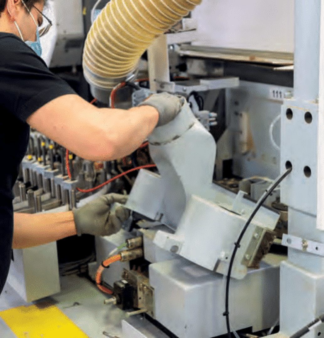 Ein Arbeiter, der Handschuhe und eine Maske trägt, bedient oder repariert eine Maschine in einem industriellen Umfeld und hantiert mit einem großen Schlauch, der mit der Maschine verbunden ist, wobei verschiedene Drähte und Komponenten sichtbar sind.