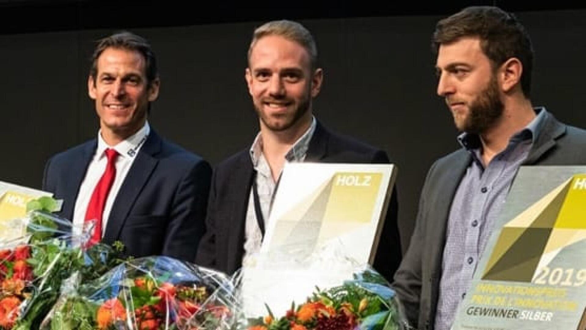 Drei Männer in förmlicher Kleidung stehen nebeneinander und halten jeweils einen Blumenstrauß und eine Plakette in der Hand. Sie lächeln, was darauf hindeutet, dass sie gerade bei einer Veranstaltung eine Anerkennung erhalten haben.