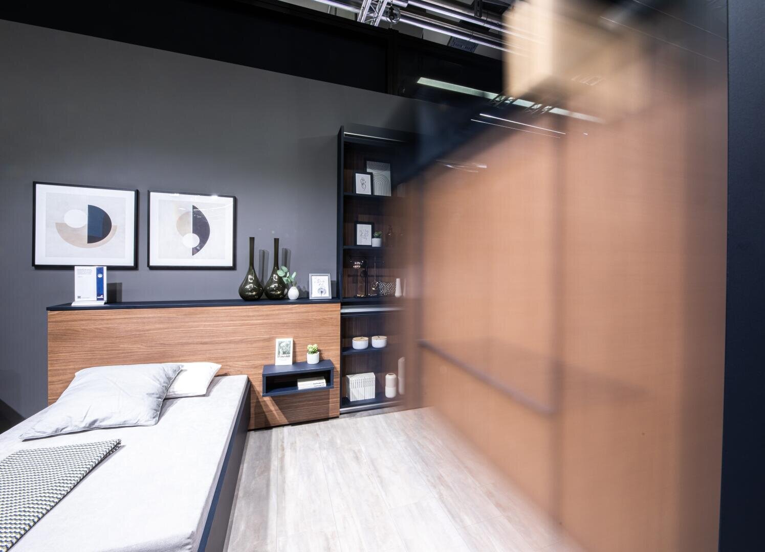 Un espace de vie moderne et minimaliste avec un canapé gris clair, un panneau latéral en bois, des vases décoratifs, un tableau abstrait encadré au mur et une étagère noire contenant des livres et de la décoration ; flou partiel sur le côté droit.