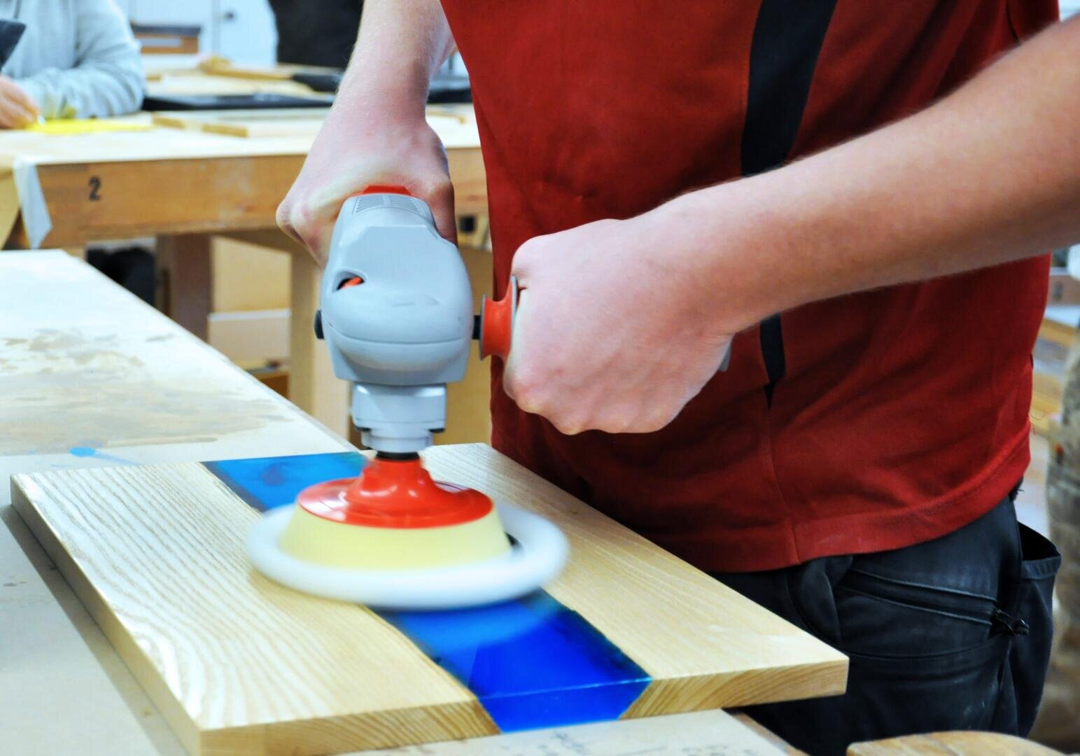 Eine Person in einem roten Hemd arbeitet in einer Werkstatt mit einer elektrischen Handpoliermaschine auf einer Holzplatte mit blauer Harzeinlage.