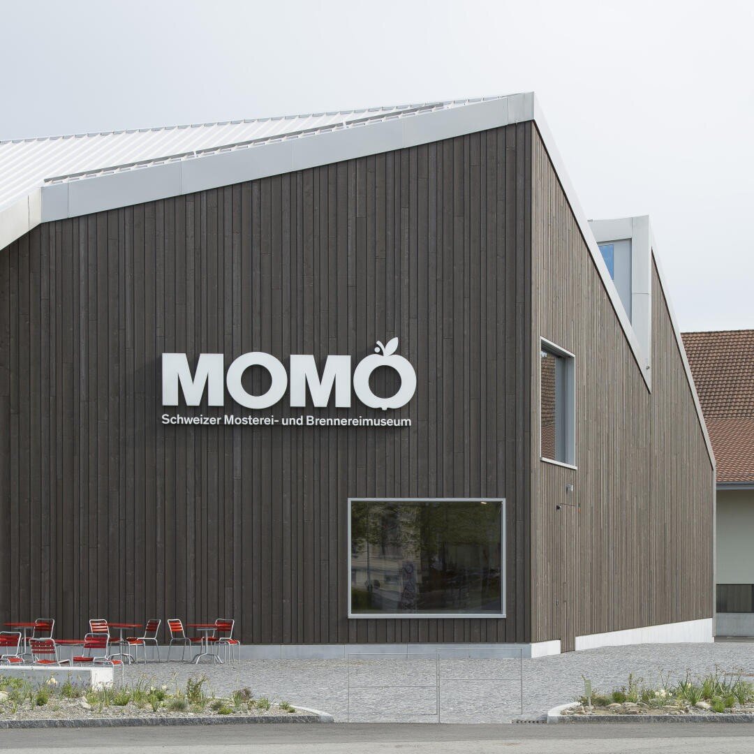 Ein modernes Gebäude mit dunkler Holzverkleidung trägt das Schild MOMO Schweizer Mosterei- und Brennereimuseum. Rote Stühle und Tische stehen draußen an einem großen Fenster.