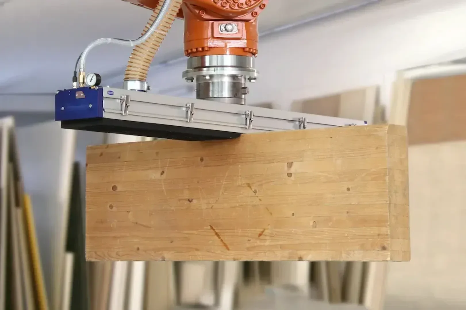 Ein Roboterarm mit einer Saugvorrichtung hebt ein großes Holzbrett in einer Werkstatt an, in der im Hintergrund verschiedene Holzplatten zu sehen sind.