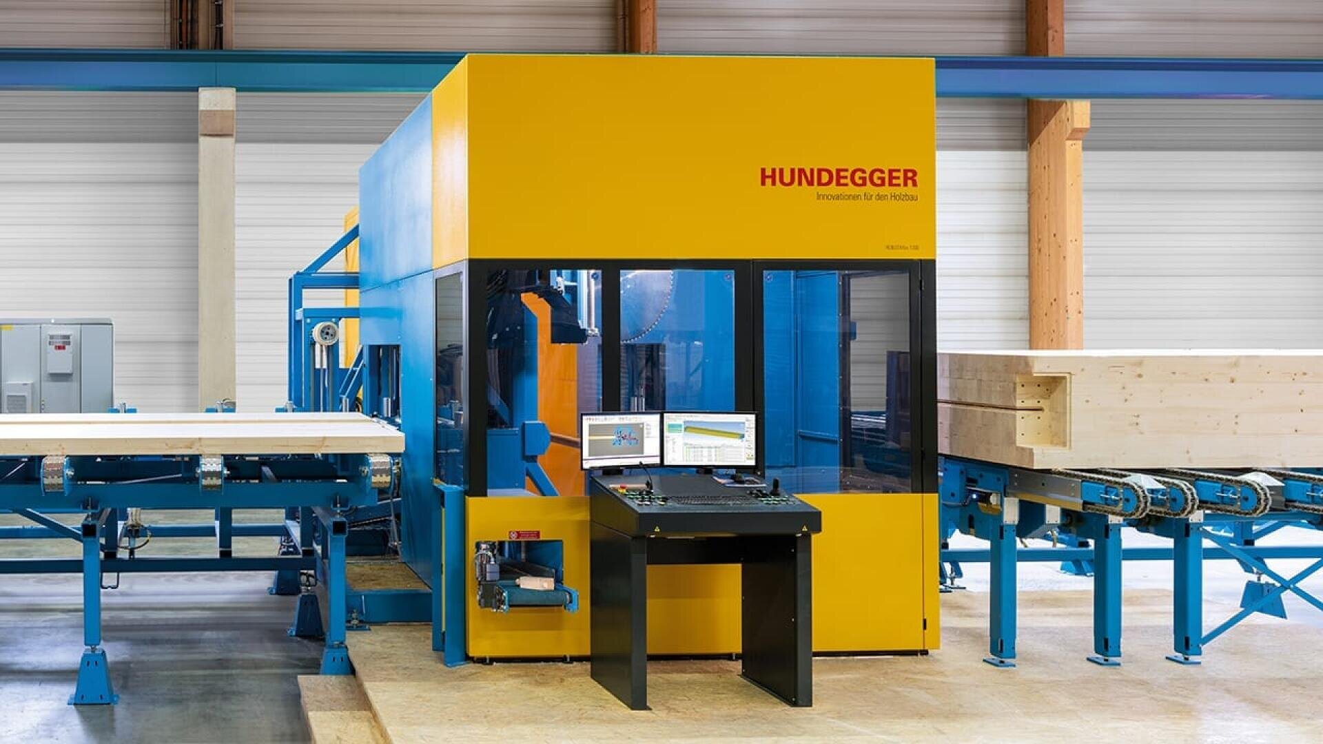 Une grande machine à bois industrielle Hundegger jaune et bleue se trouve dans une usine. Devant elle se trouvent deux écrans d'ordinateur sur un bureau noir, avec des poutres de bois sur des rouleaux qui entrent et sortent de la machine.