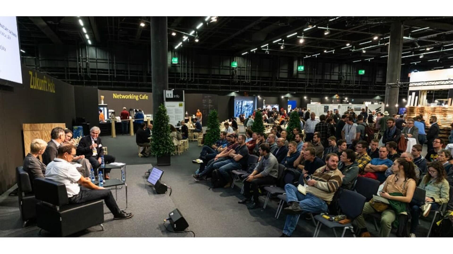 Un grand groupe de personnes est assis face à un groupe d'orateurs lors d'une conférence dans un lieu spacieux et moderne. Des panneaux tels que Networking Café sont visibles et les participants écoutent attentivement.