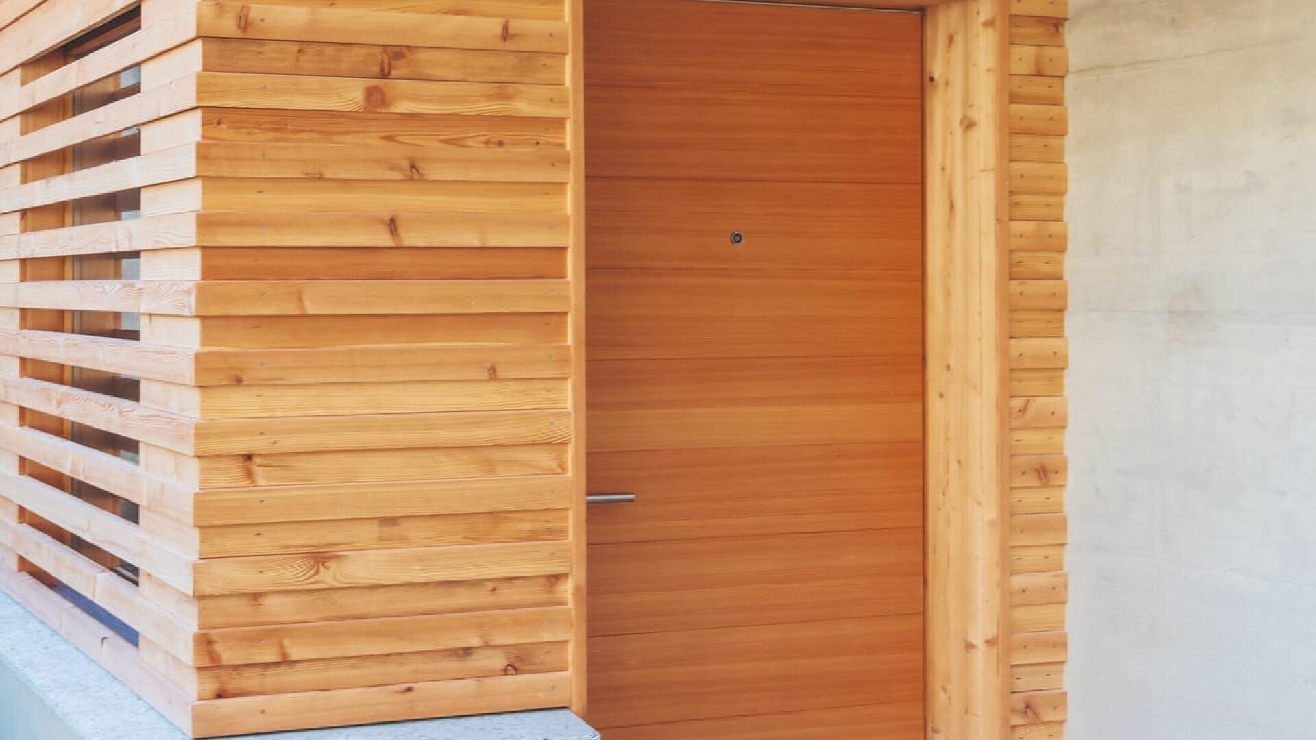Une porte moderne en bois encastrée dans un mur avec des lattes horizontales en bois, à côté d'une surface en béton lisse. Le design présente des lignes épurées et une esthétique minimaliste.