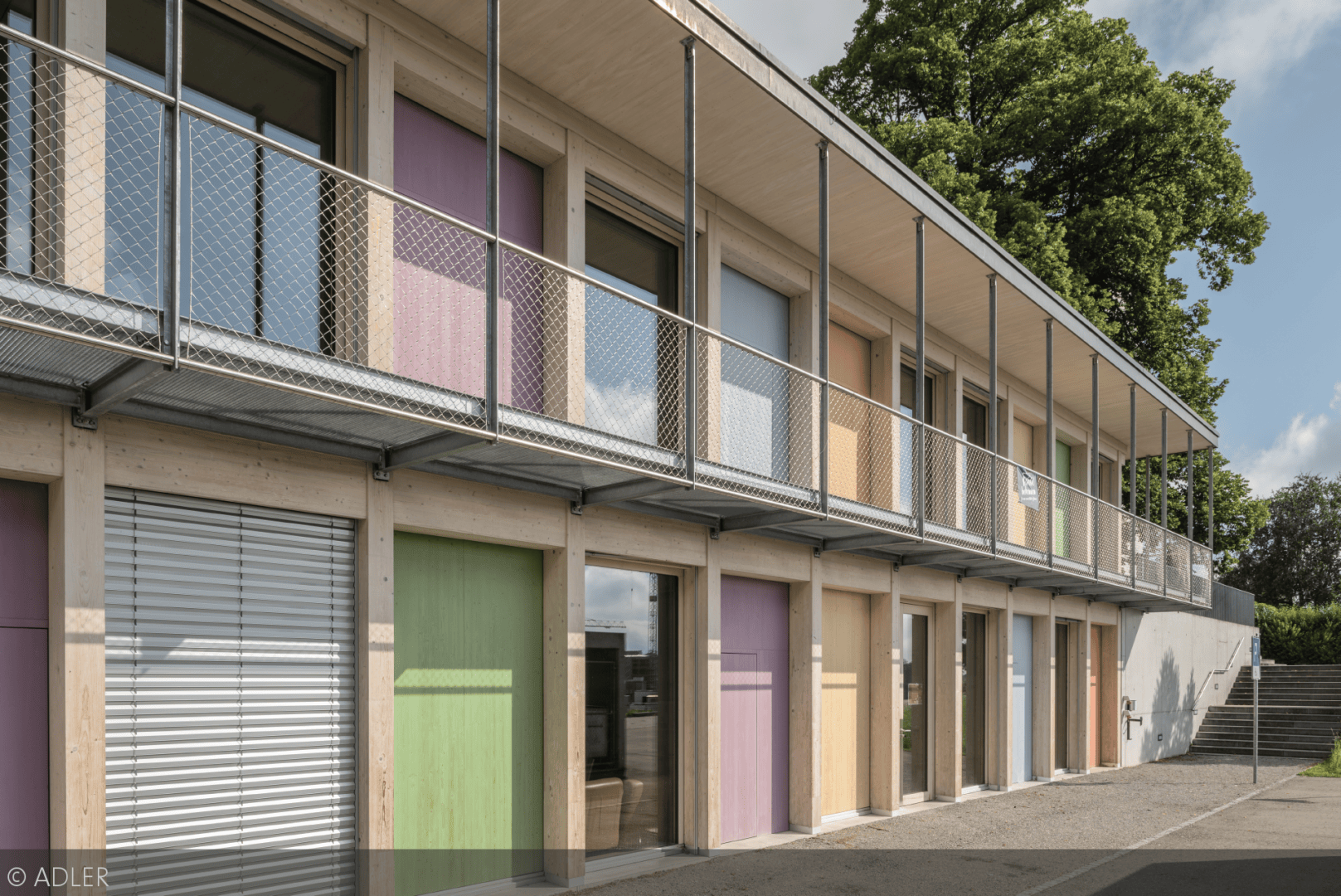 Ein zweistöckiges modernes Gebäude mit pastellfarbenen Türen im Erdgeschoss, großen Fenstern, einem Metallbalkon mit Netzgeländer im Obergeschoss und Bäumen im Hintergrund unter einem teilweise bewölkten Himmel.