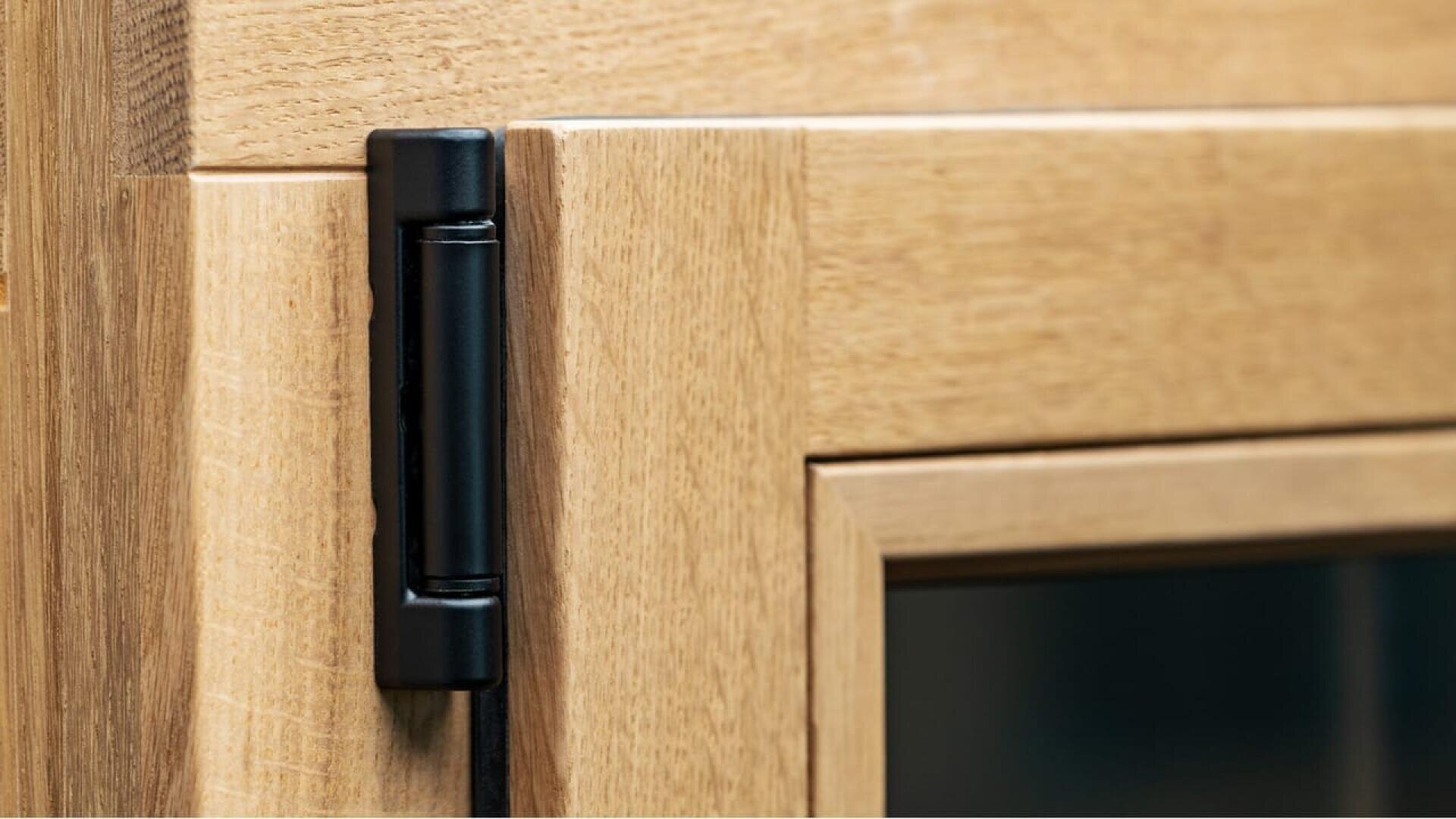 Gros plan d'un cadre de fenêtre en bois sur lequel est fixée une charnière en métal noir, montrant la texture du bois clair et une partie de la vitre.
