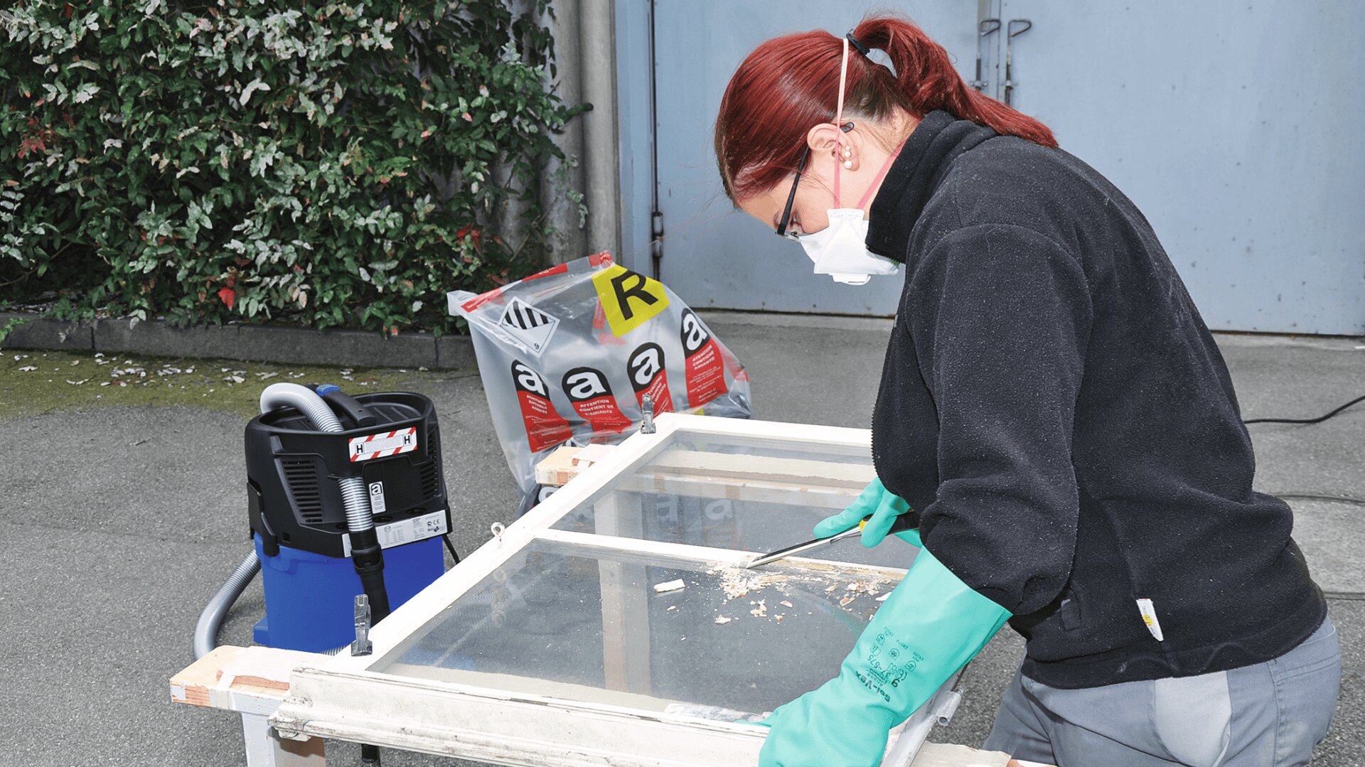 Eine Person, die eine Maske, Handschuhe und Schutzkleidung trägt, kratzt im Freien die Farbe von einem Fensterrahmen ab. In der Nähe befinden sich Säcke mit Materialien und ein Staubsauger, im Hintergrund ist Grünzeug zu sehen.