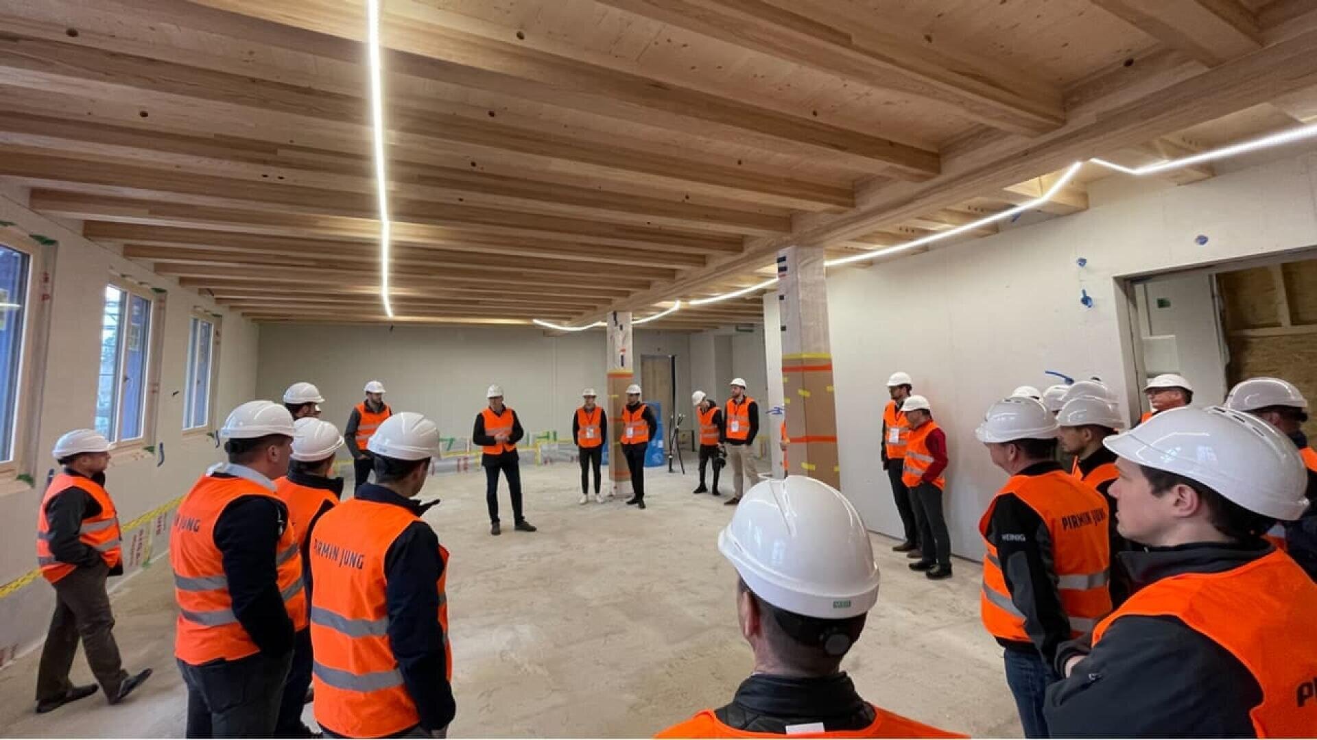 Un groupe de personnes portant des casques blancs et des gilets de sécurité orange se tient en cercle à l'intérieur d'une grande pièce inachevée avec des poutres en bois et des murs blancs, probablement sur un chantier de construction.
