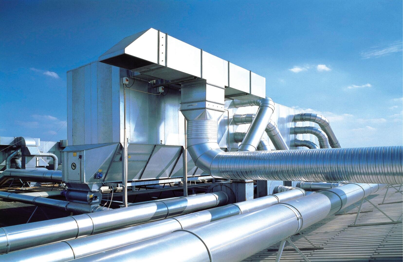 Großes industrielles HVAC-System mit silbernen Metallkanälen, Rohren und Lüftungseinheiten, das auf einem Dach vor einem klaren blauen Himmel installiert ist.