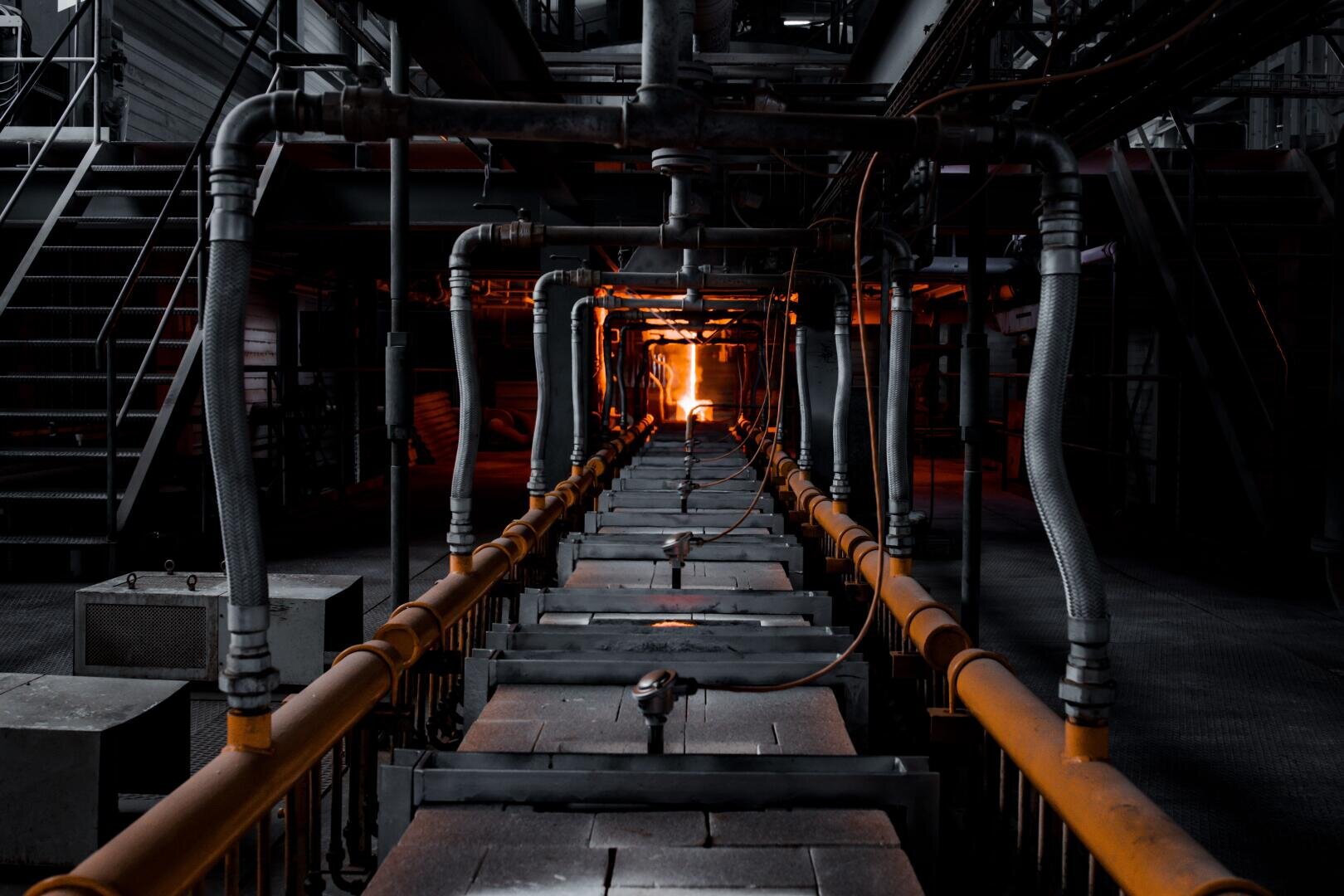 Innenansicht einer Industrieanlage mit Metallrohren und Maschinen, die zu einem glänzenden Ofen am Ende eines langen Korridors führen und ein intensives orangefarbenes Licht erzeugen.