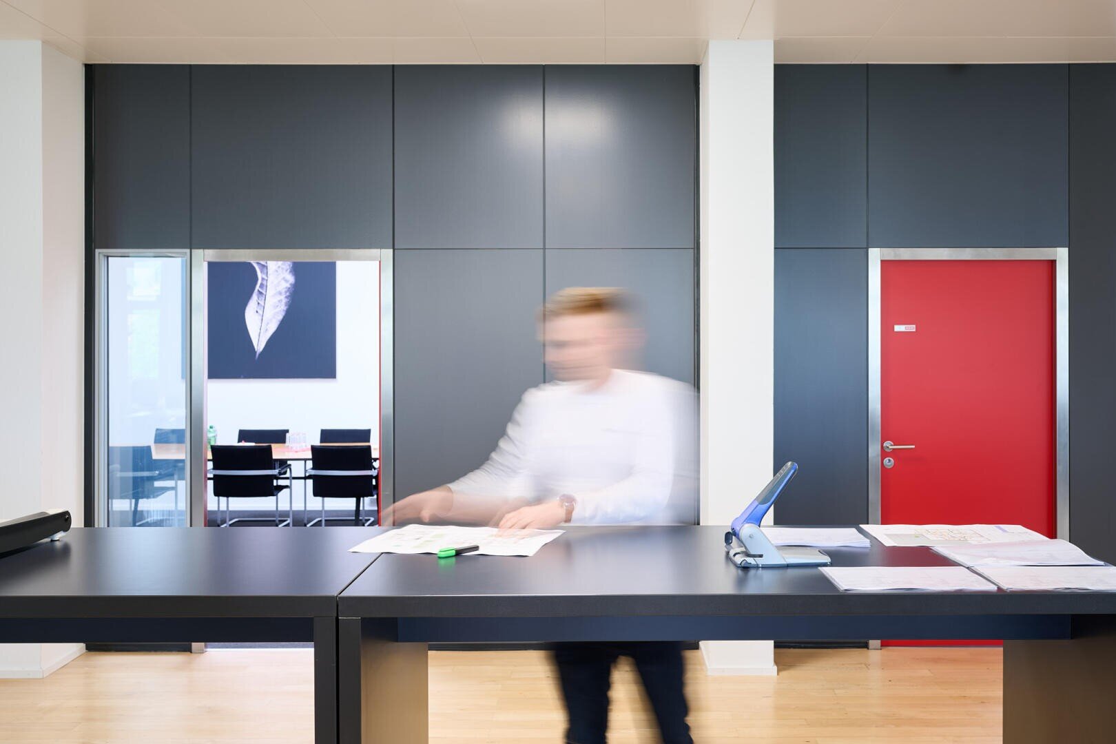 Une personne en mouvement se tient devant un comptoir d'accueil de bureau moderne avec des panneaux sombres, une porte rouge et une salle de conférence où l'on peut voir un tableau en arrière-plan. Le visage de la personne est flou en raison du mouvement.