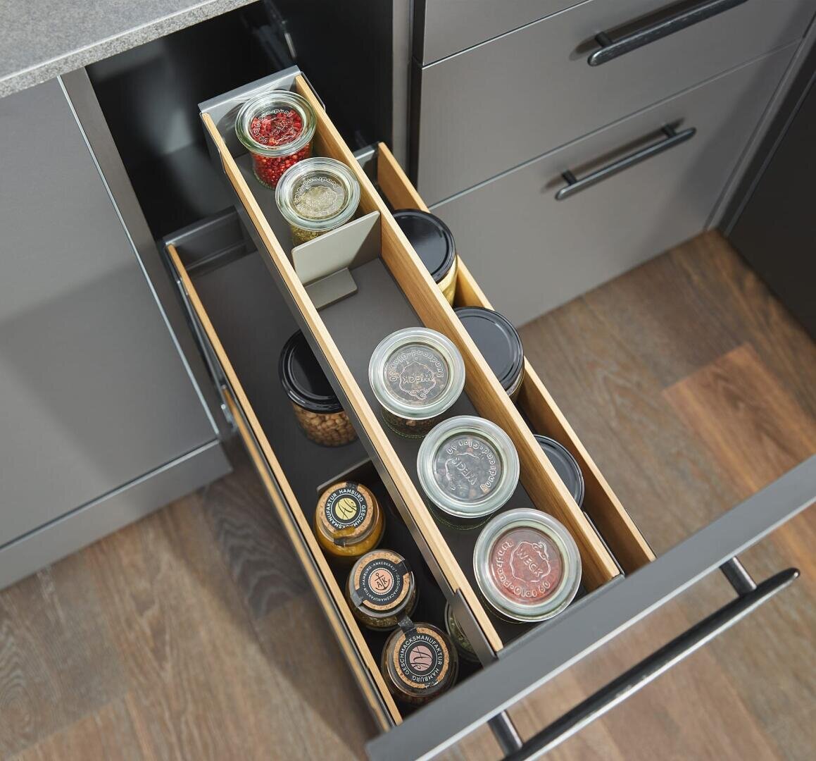 Eine Küchenschublade mit hölzernen Trennwänden sorgt für Ordnung in den Gewürzdosen und Behältern in einem modernen grauen Schrank mit dunklen Griffen und einem holzgemusterten Boden.