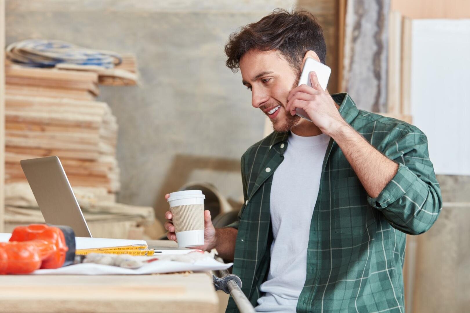 Un homme vêtu d'une chemise à carreaux verts tient une tasse de café à la main et téléphone tout en travaillant sur un ordinateur portable dans un atelier de menuiserie, entouré d'outils et de planches de bois.