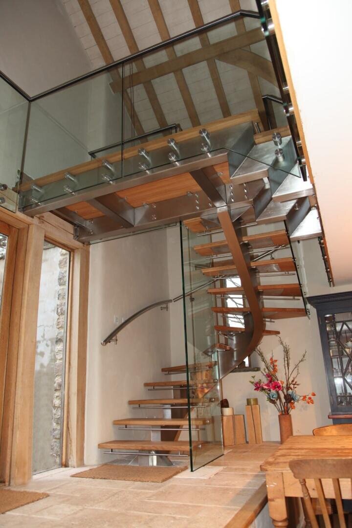 Eine moderne schwebende Holztreppe mit Glasgeländer schlängelt sich in einem hellen Raum mit freiliegenden Balken, großen Fenstern und einem Esstisch aus Holz mit Blumen darauf nach oben.