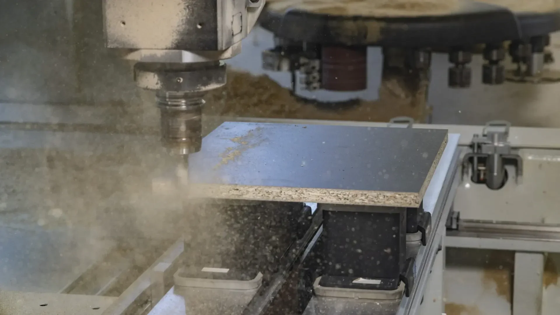 Eine CNC-Fräsmaschine schneidet in ein Stück Holzwerkstoff, wobei Sägespäne und Partikel entstehen. Das Werkstück ist auf dem Maschinenbett befestigt, und das rotierende Schneidwerkzeug ist in Aktion.
