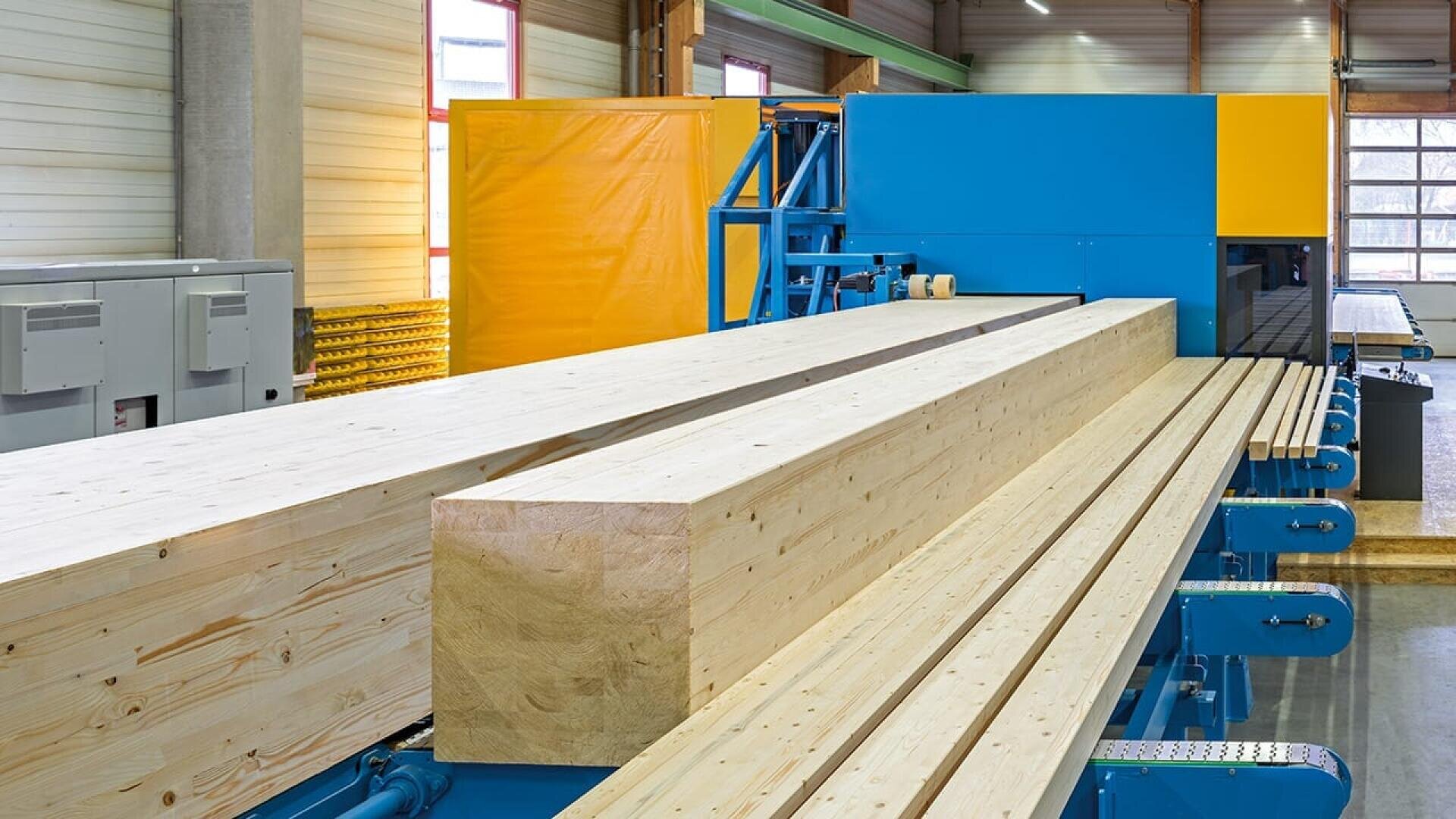 De grandes poutres en bois sur un système de convoyeur bleu à l'intérieur d'une installation industrielle moderne et bien éclairée. L'équipement et les murs sont propres, avec des panneaux jaunes et orange et des fenêtres qui laissent entrer la lumière naturelle.