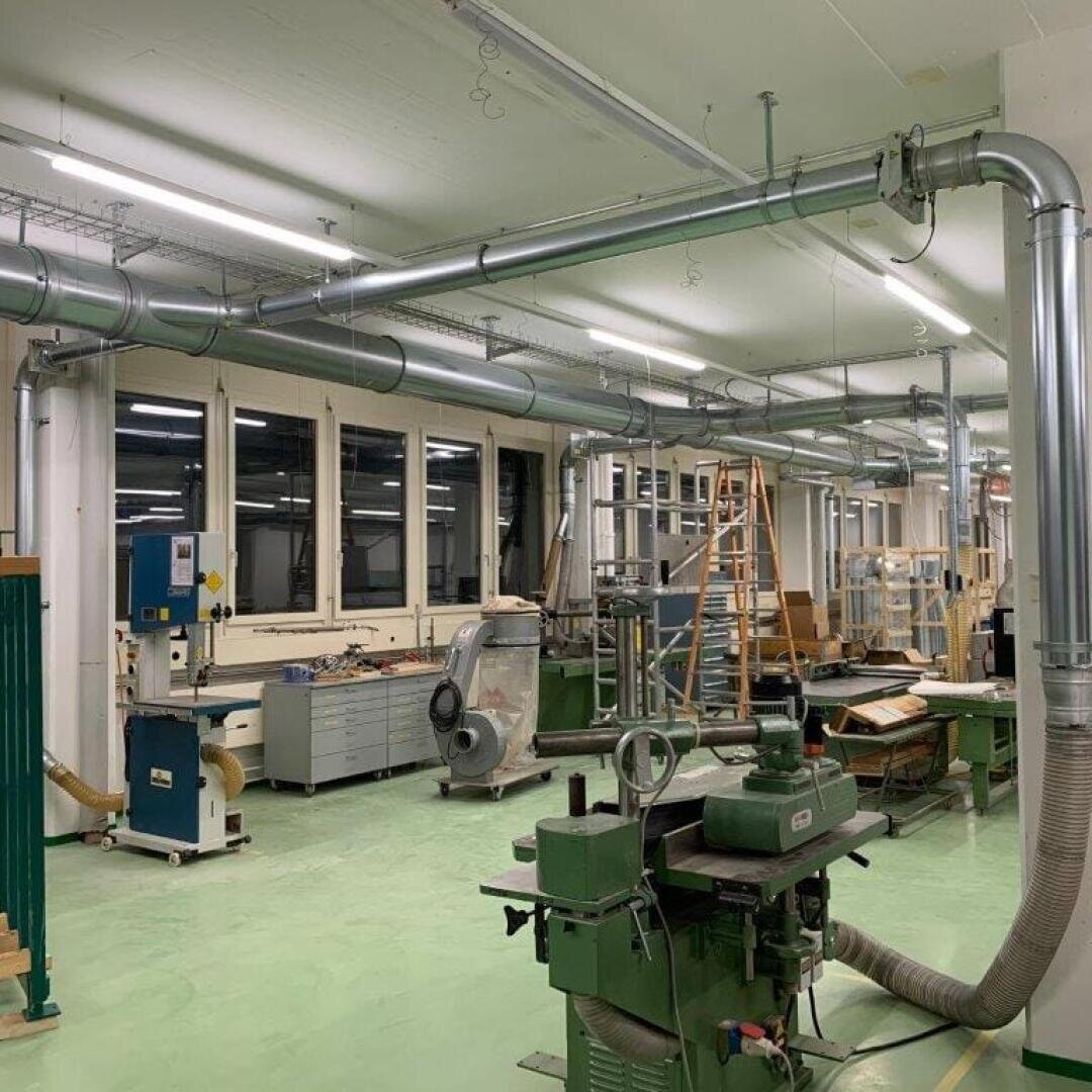 Eine geräumige, gut beleuchtete Werkstatt mit verschiedenen Holzbearbeitungsmaschinen, Rohren an der Decke, Werkbänken und Werkzeugen. Große Fenster säumen eine Wand, und eine Leiter steht in der Nähe der Rückseite des Raumes.