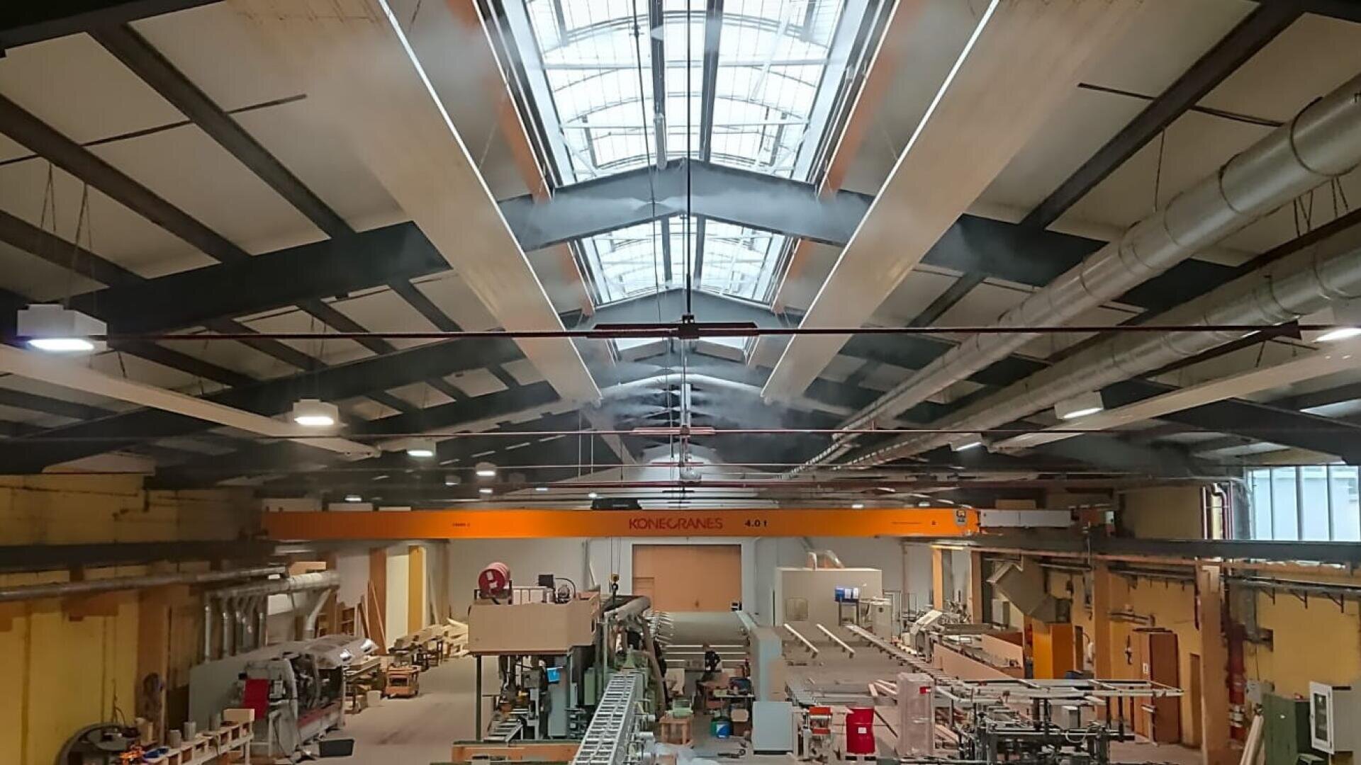 Großes Industrielager mit hohen Decken, Oberlichtern, freiliegenden Balken und sichtbaren Maschinen, Arbeitsplätzen und Geräten; ein gelber Brückenkran mit der Aufschrift Konecranes überspannt den Raum.