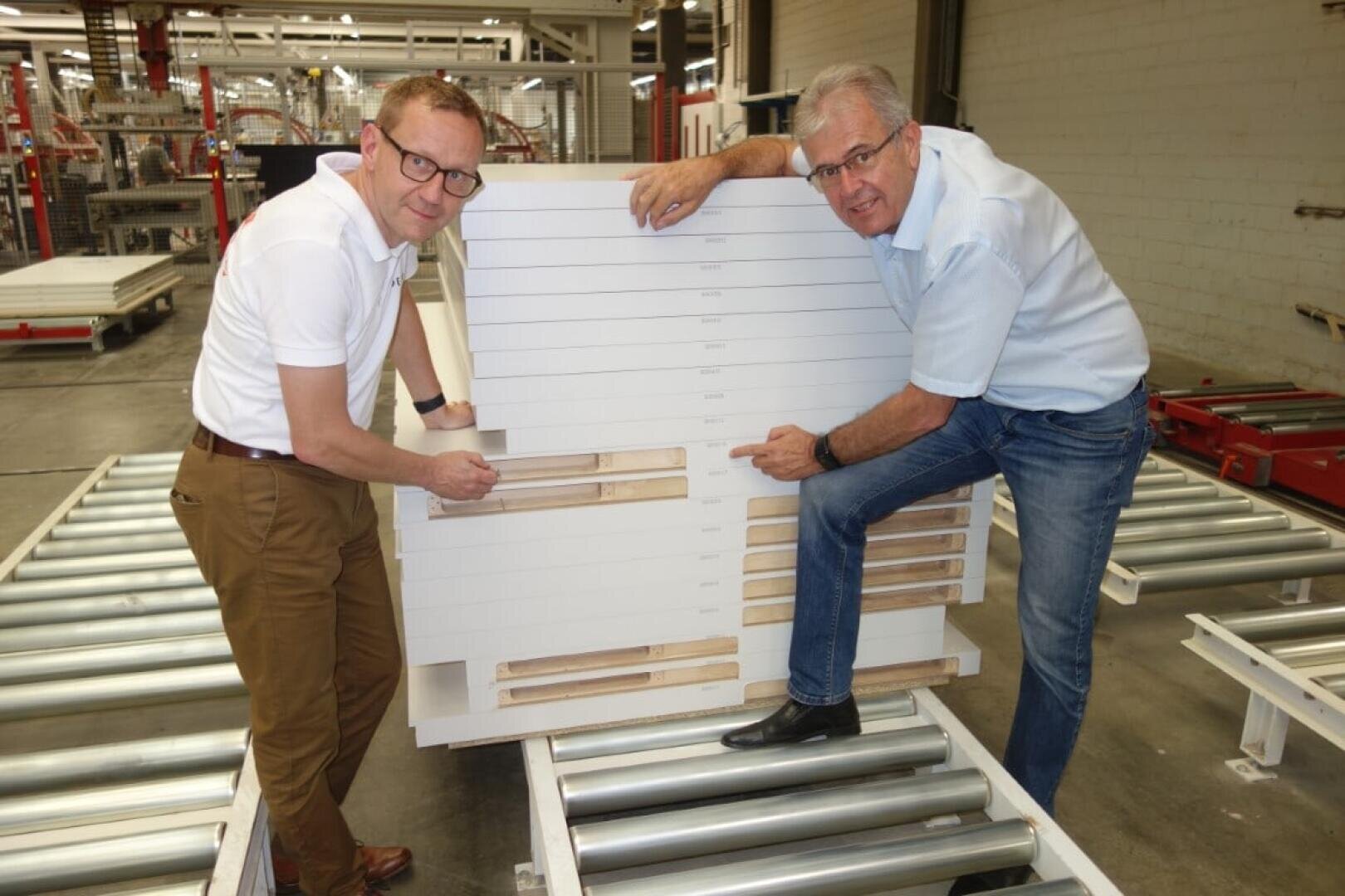 Zwei Männer mit Hemden und Brillen stehen in einem Lagerhaus und zeigen auf eine große weiße Platte mit Holzlatten, die auf Metallrollen steht. Sie lächeln und demonstrieren die Eigenschaften der Platte.
