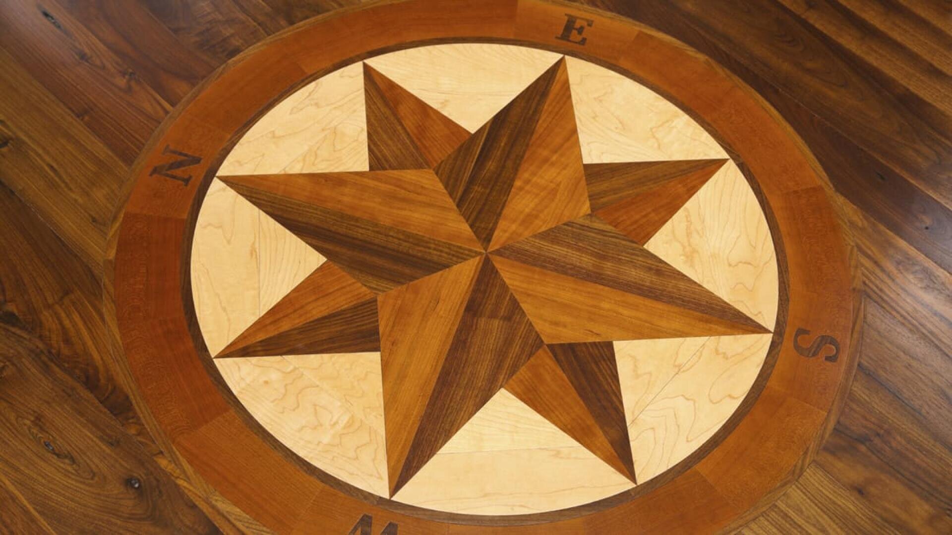 Eine hölzerne Kompassrose mit acht Punkten und den Himmelsrichtungen (N, E, S, W), die in einen polierten Holzboden eingelassen ist. Das Design verwendet kontrastierende Holztöne für den visuellen Effekt.