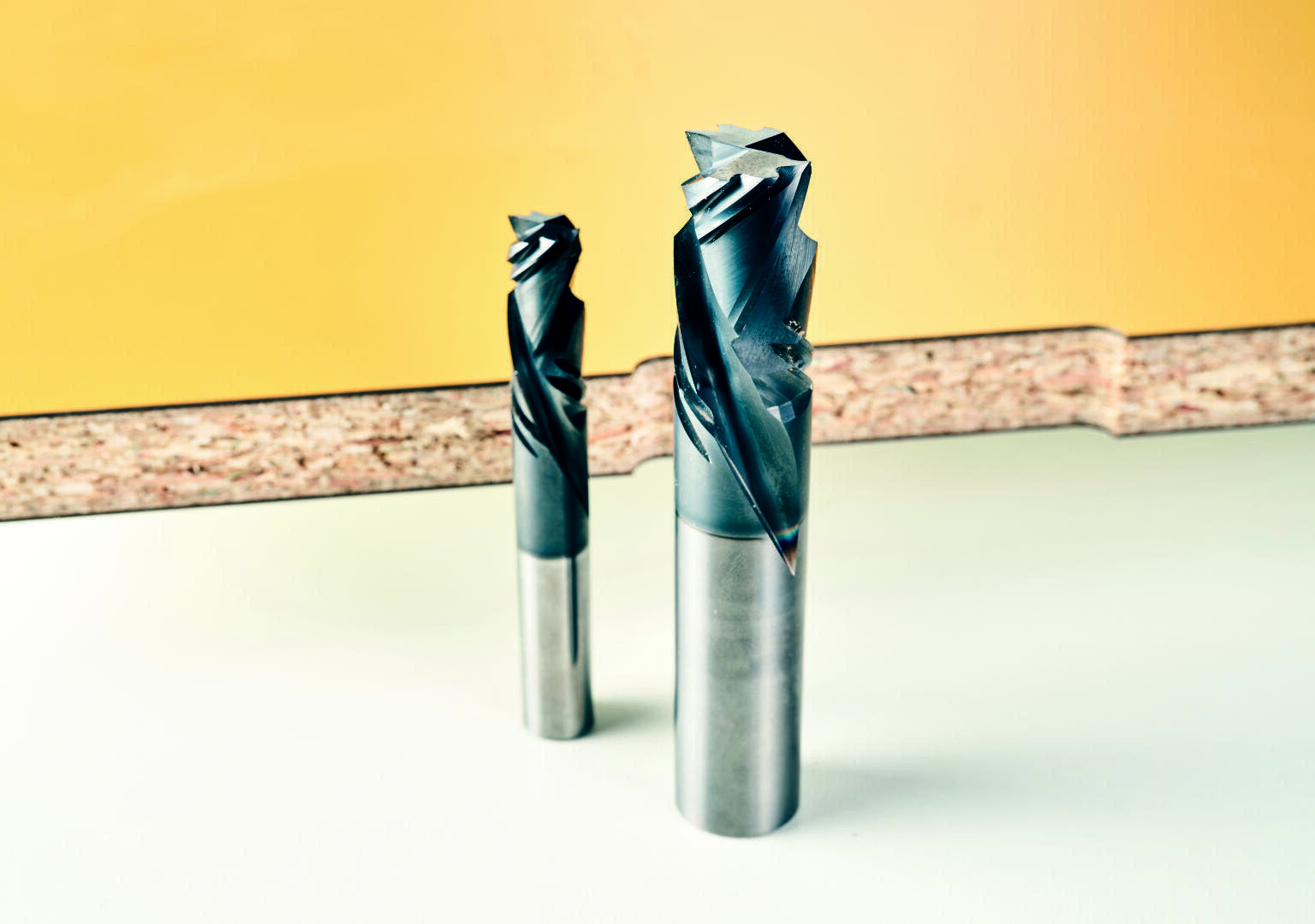 Zwei Metallfräser stehen aufrecht auf einer hellen Fläche mit einer gelben Wand und einem Streifen aus strukturiertem Material im Hintergrund.