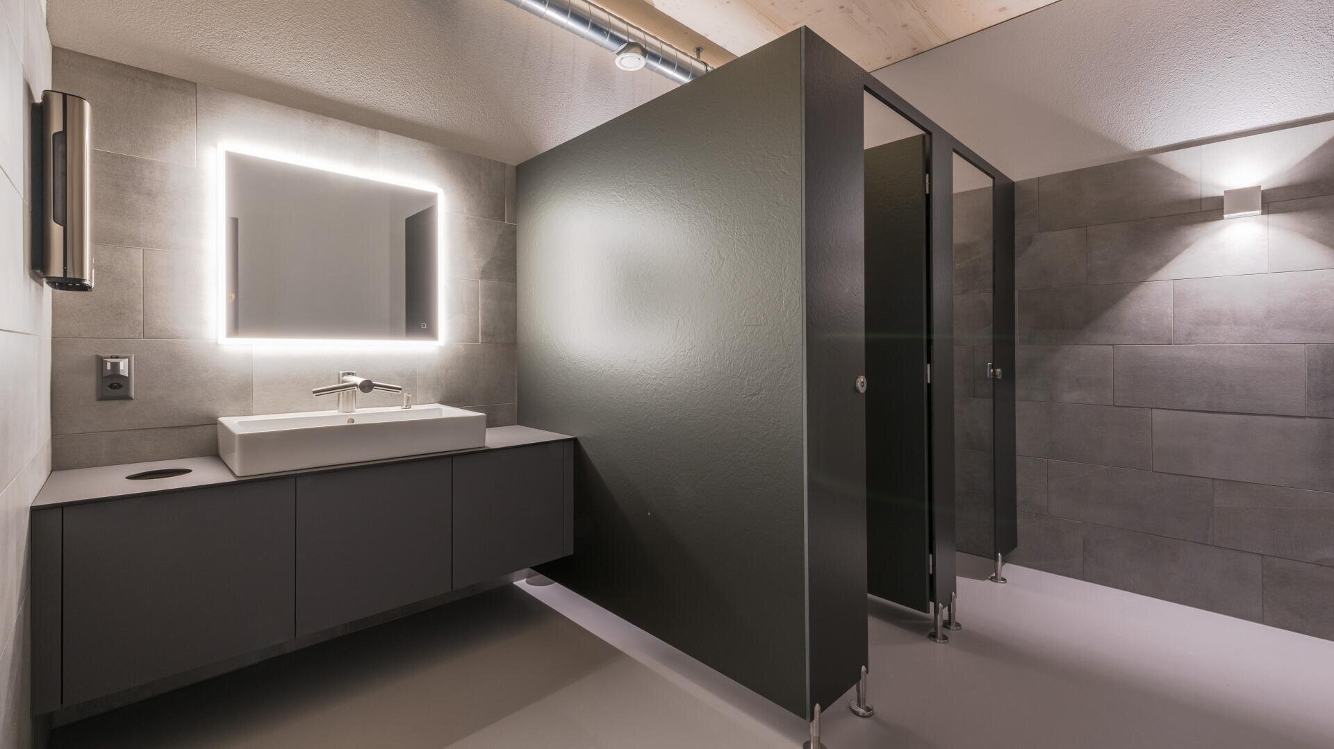 Eine moderne öffentliche Toilette mit grauen Wänden, einem großen beleuchteten Spiegel über einem Waschbecken und einem Tresen sowie zwei dunkel gefärbten Toilettenkabinen, die durch Trennwände voneinander getrennt sind.