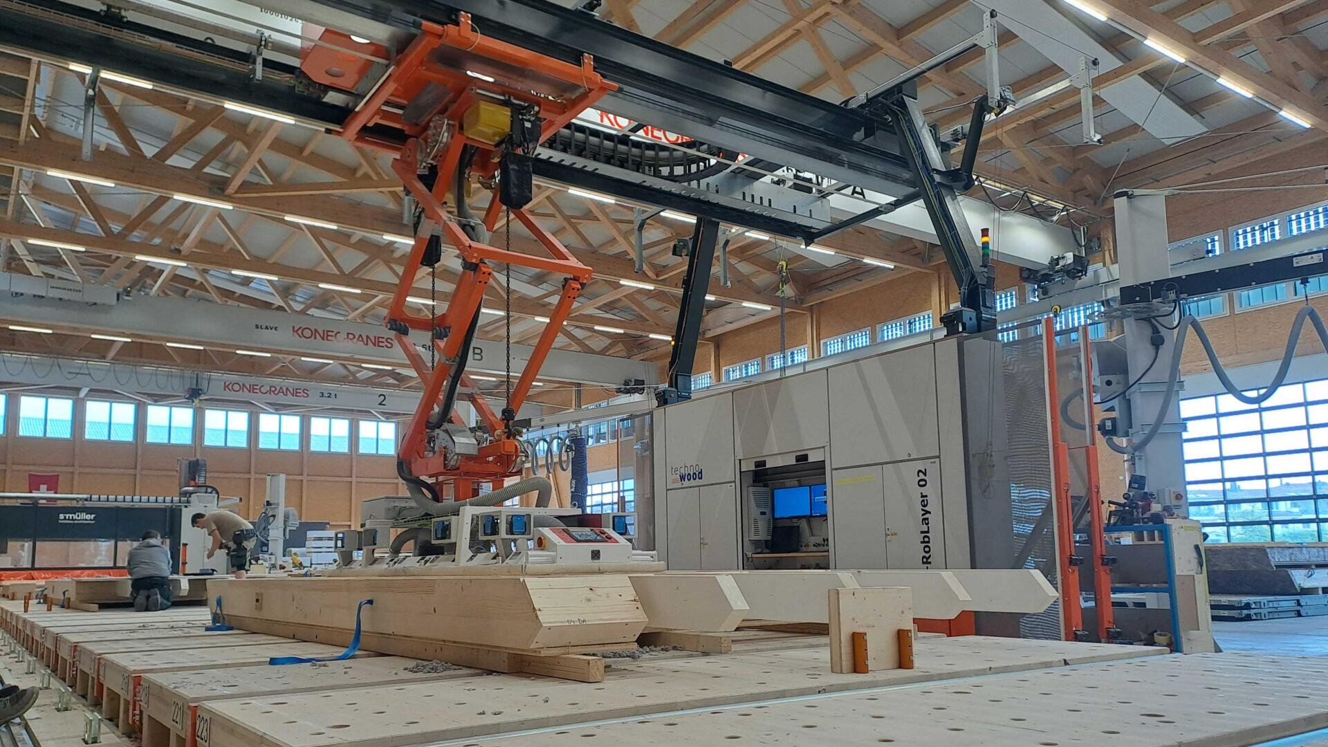 Ein großer Industrieroboterarm montiert Holzbalken in einer geräumigen, modernen Fabrik mit hohen Decken und reichlich natürlichem Licht aus großen Fenstern. Verschiedene Maschinen und Anlagen sind im Hintergrund zu sehen.